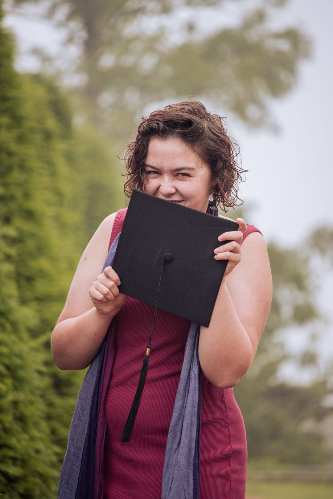 Smiling graduate playfully hides behind black graduation cap in lush green garden setting.