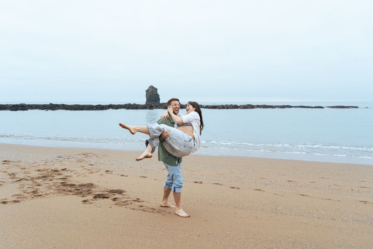 Man lifting and spinning pregnant partner joyfully on sandy beach by the ocean.