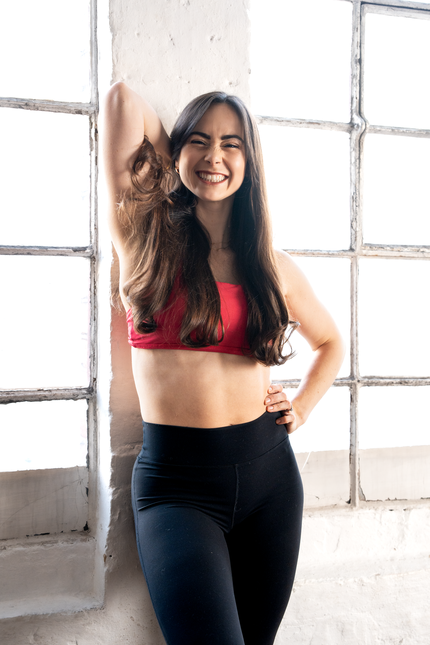 Laughing brunette woman in red sports bra and black leggings posing playfully by window.