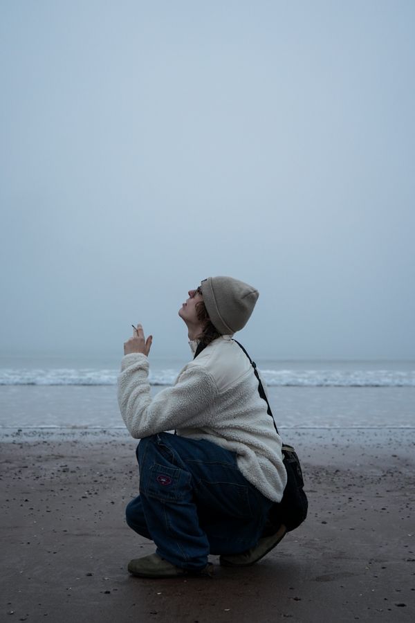 Person crouching on misty beach in white fleece and beanie, looking up while holding cigarette.