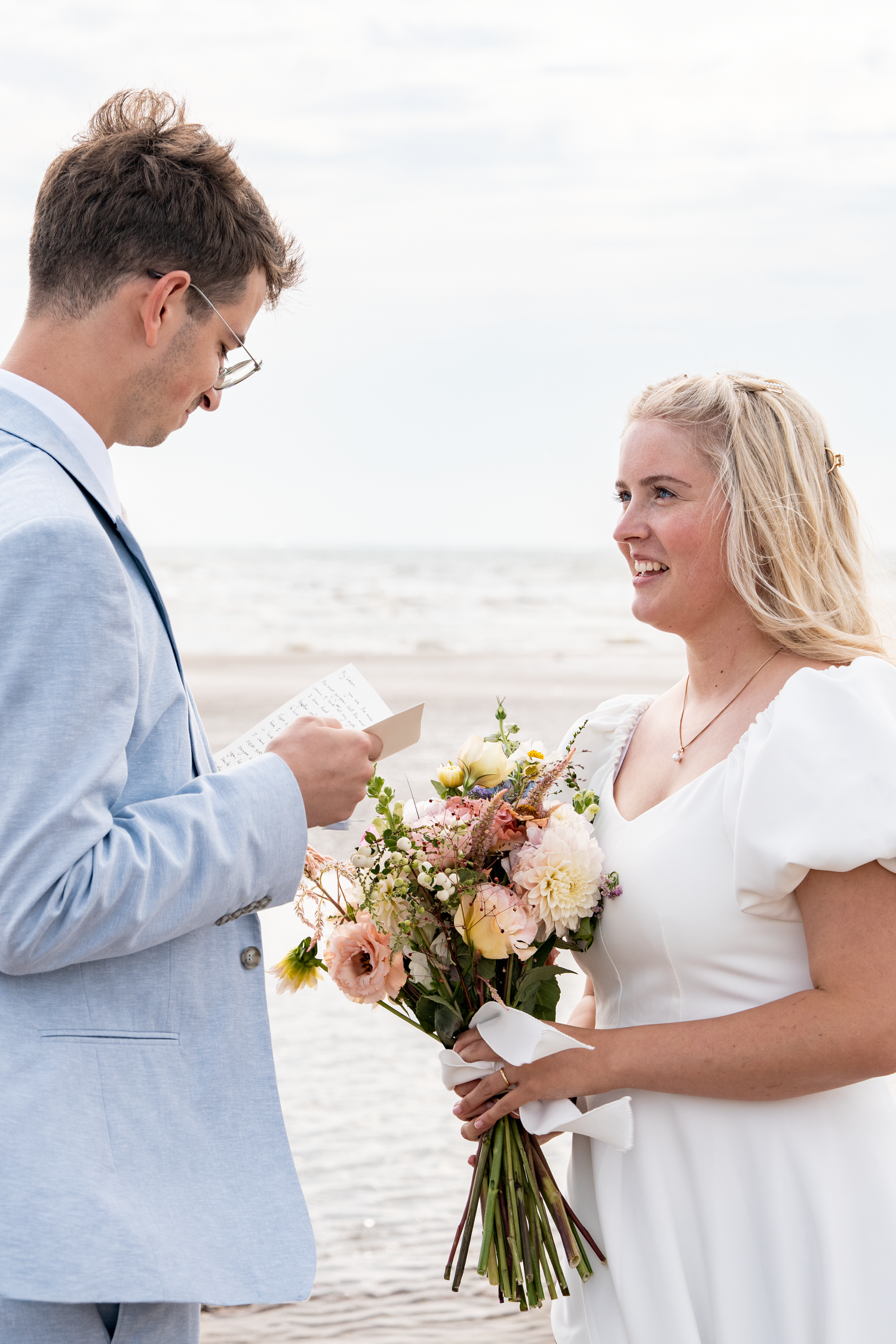 Groom in blue suit reads vows to smiling blonde bride holding floral bouquet during beach wedding ceremony.