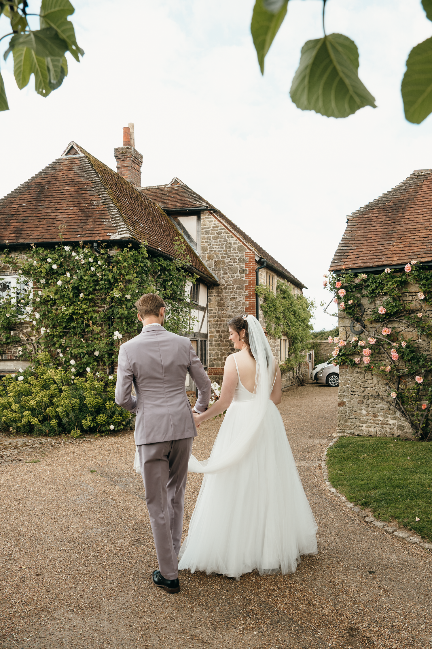 A bride and groom walk hand in hand toward a charming stone cottage surrounded by roses on their wedding day.