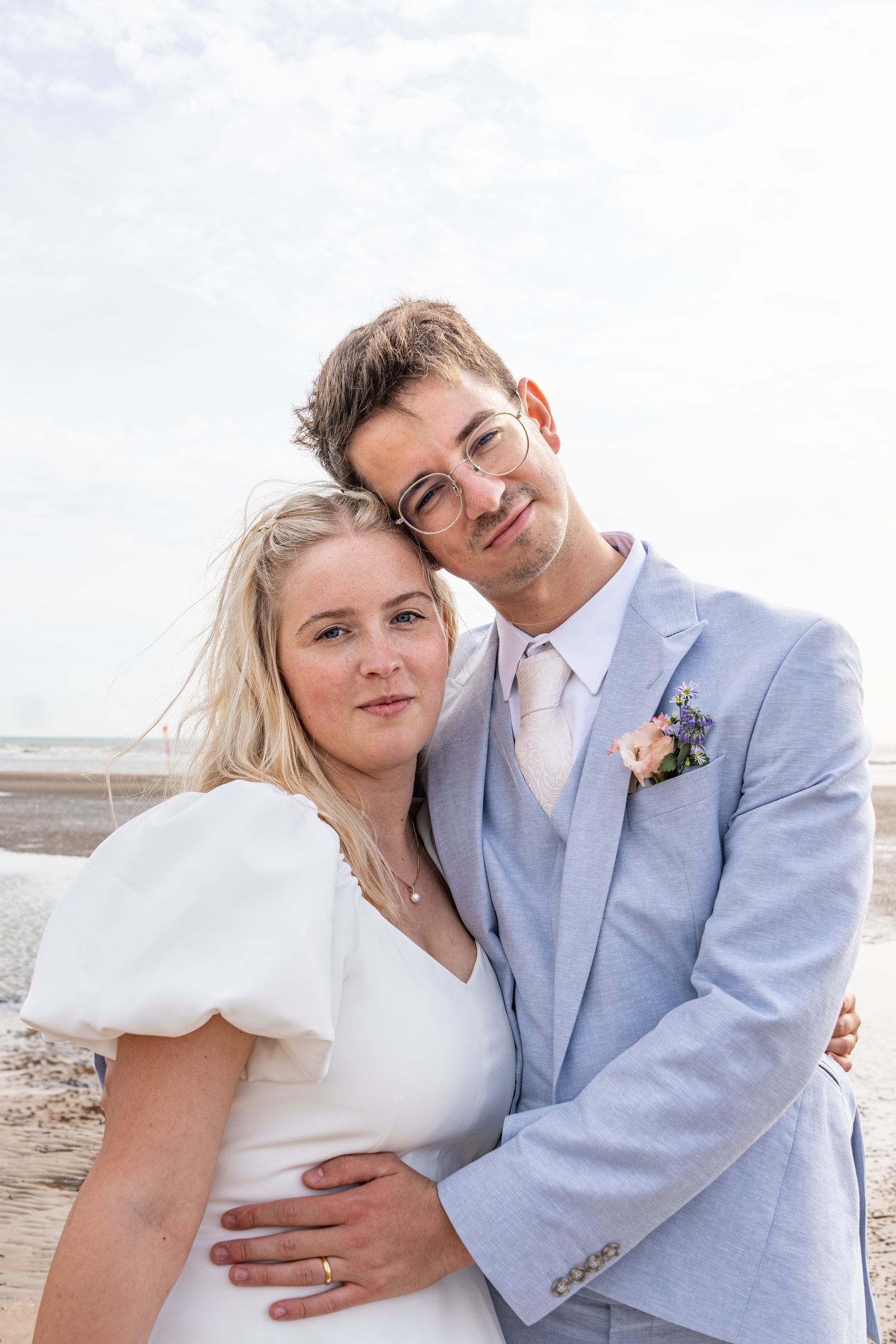 Smiling bride and groom in light blue suit embracing on sandy beach after wedding.