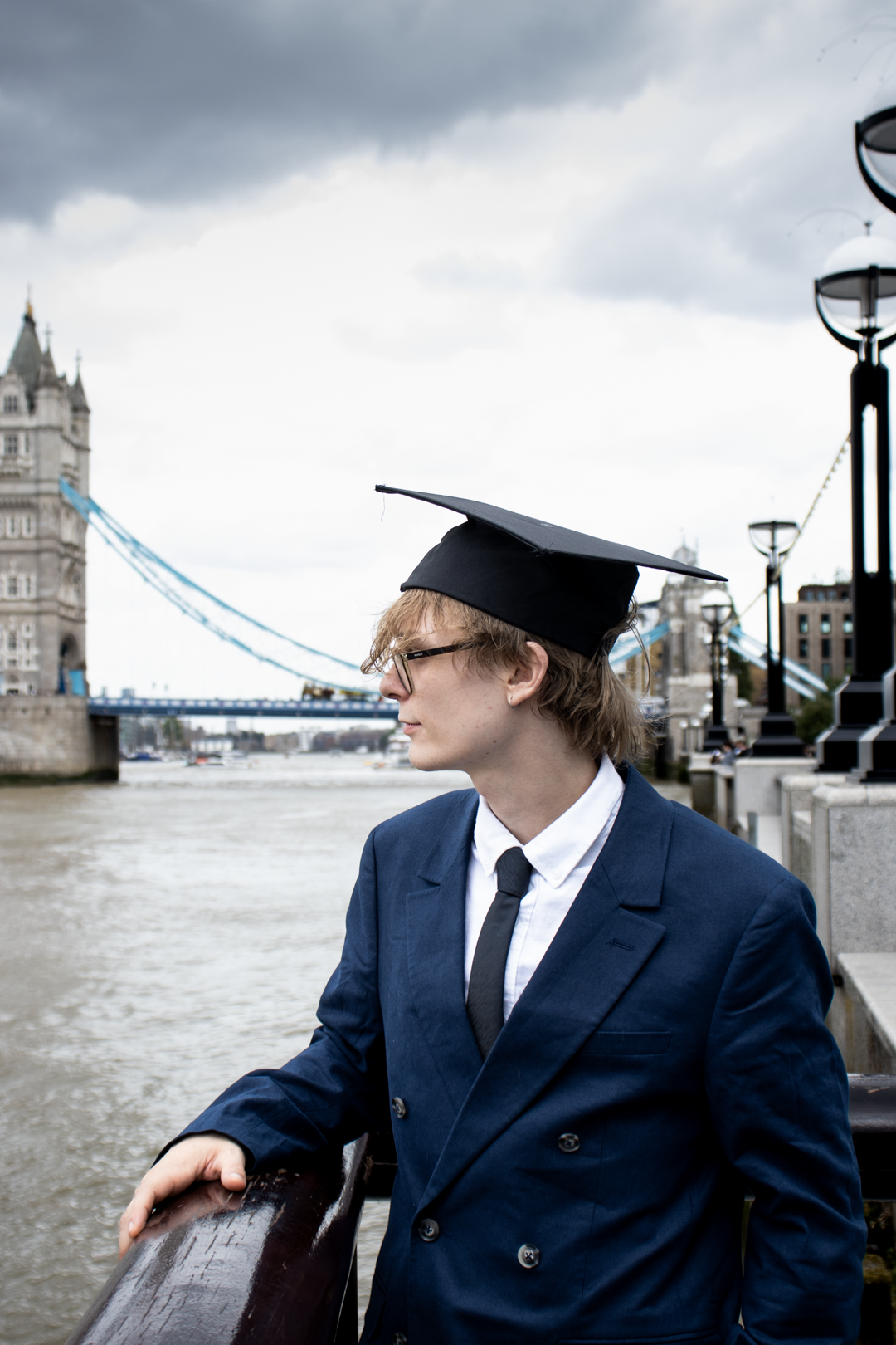 A young graduate in a navy suit and graduation cap gazes thoughtfully with Tower Bridge visible behind him.