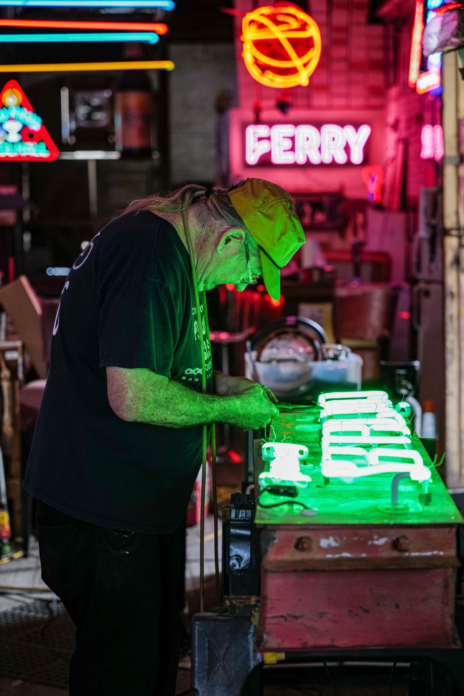 Neon sign maker working on a glowing green sign inside a studio filled with colorful neon lights.