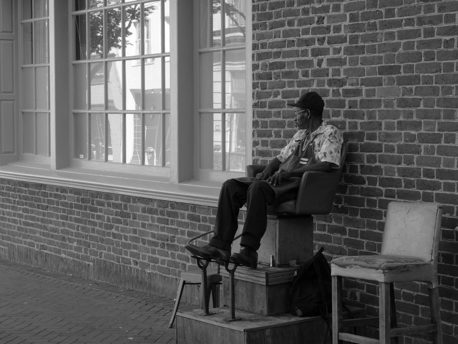Shoe shine man sitting in a chair outside a brick building in a small town setting.