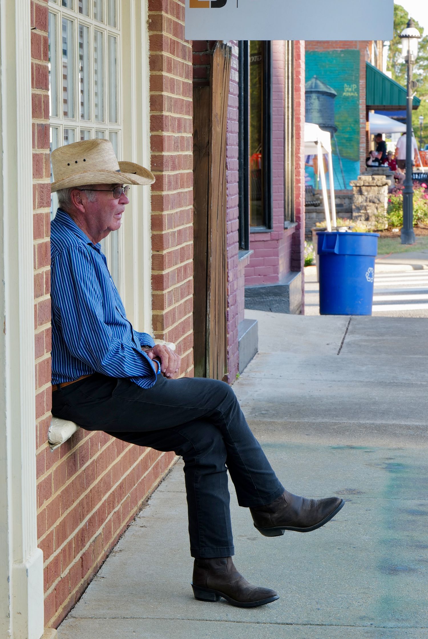 A man in a cowboy hat rests against a brick wall along a quiet sidewalk in a rural Virginia town.
