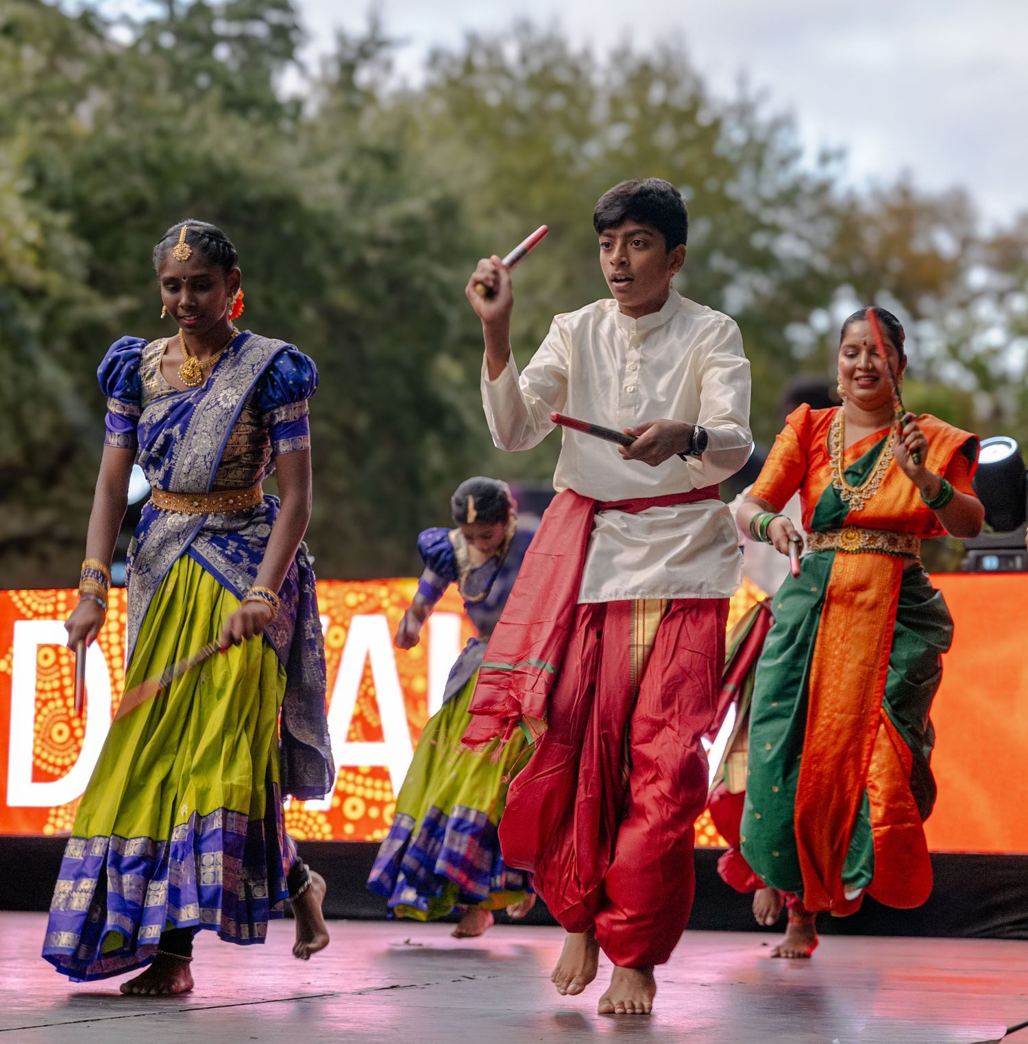 Dancers in vibrant traditional Indian attire perform a colorful dance routine on an outdoor stage during a cultural event.