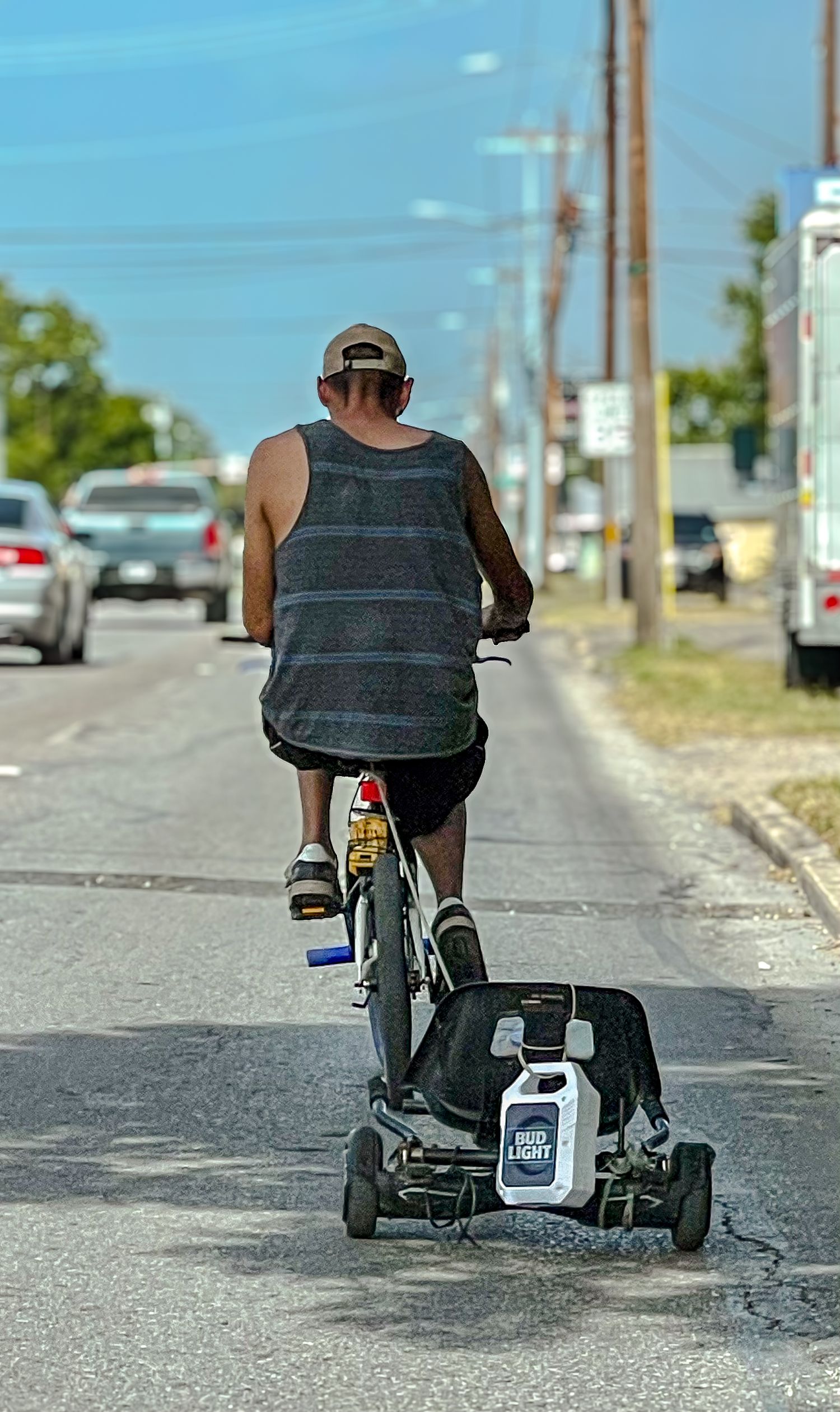 Man on a bicycle towing a makeshift cart along a busy road in an urban setting.
