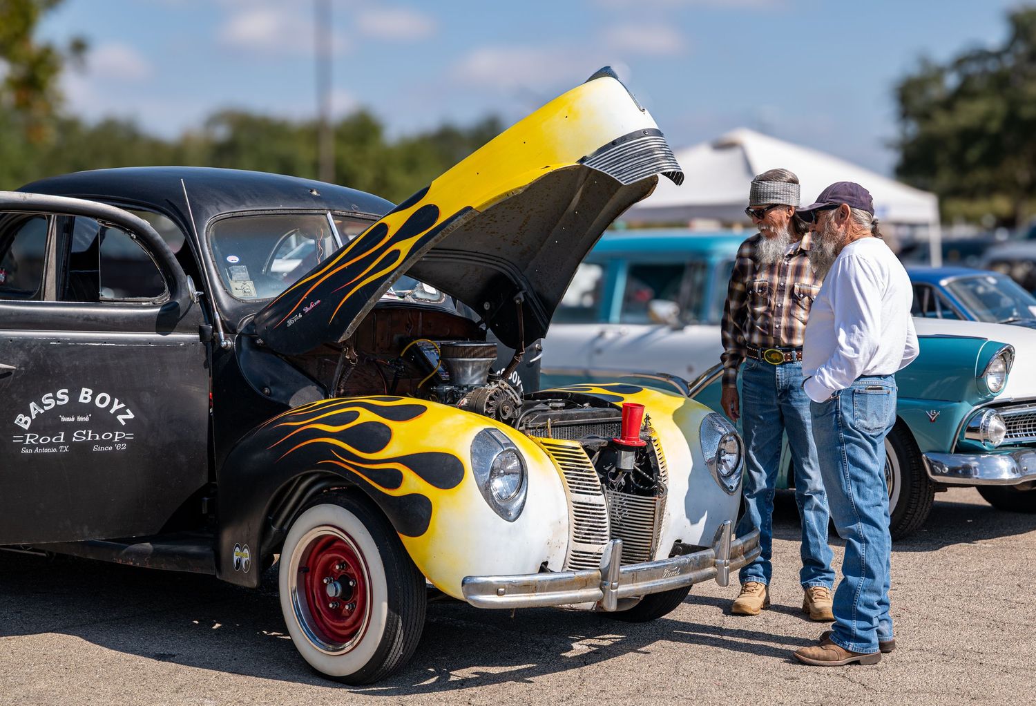Two men looking at a vintage hot rod with flame paint and open hood at a classic car show.