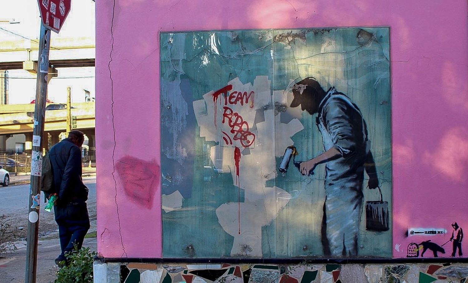 Street art mural in the style of Banksy showing a man painting graffiti on a pink wall in an urban setting.