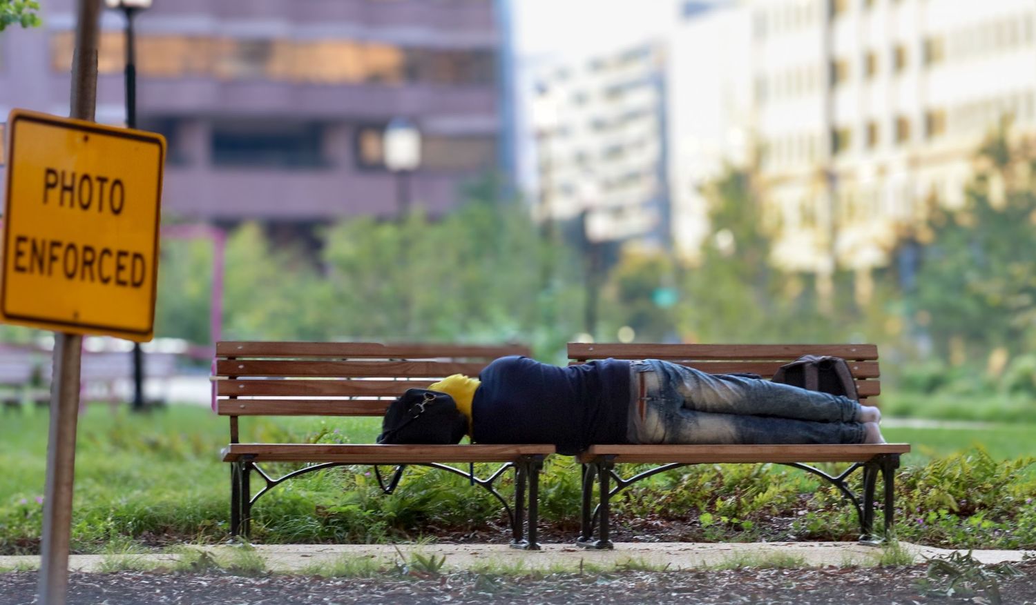 Person lying on a park bench near a “photo enforced” sign in an urban city setting.