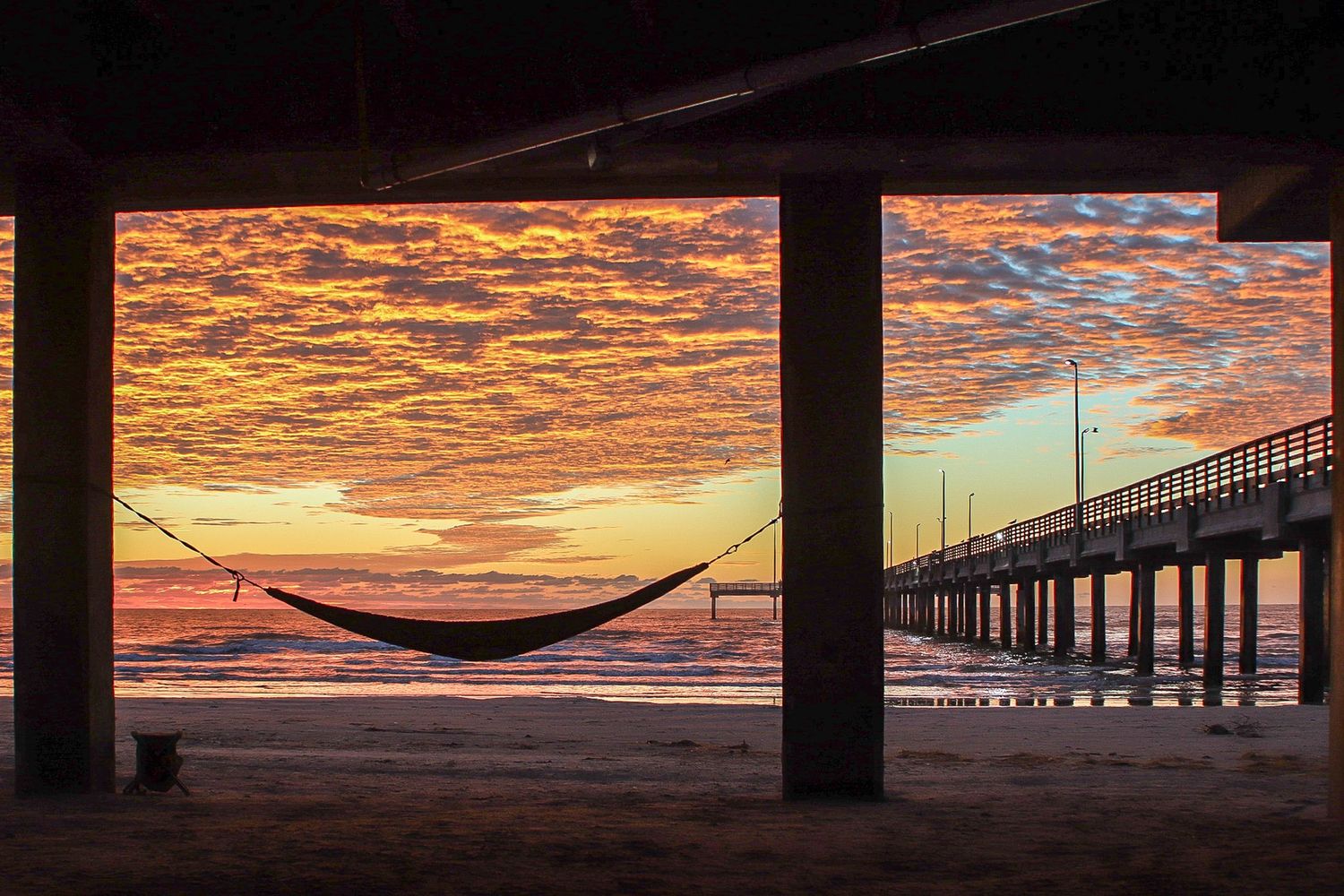 Hammock hanging beneath a beach structure at sunrise along the Texas coast, with a pier extending over the ocean and dramatic cloud formatio