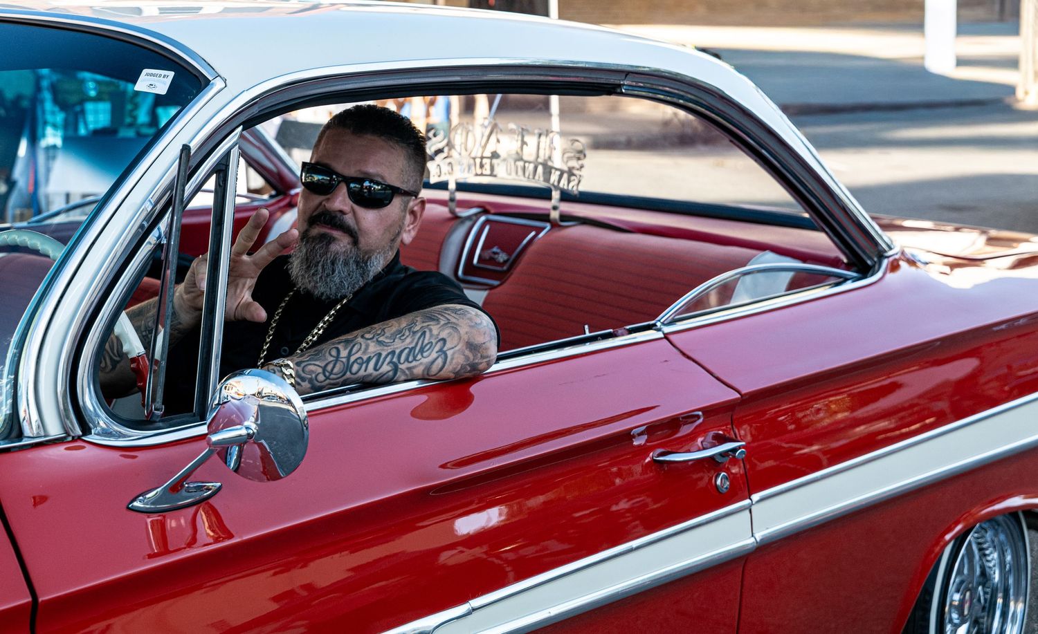 Man sitting in a vintage red car giving a hand gesture, capturing a moment of personality and car culture in a community setting.