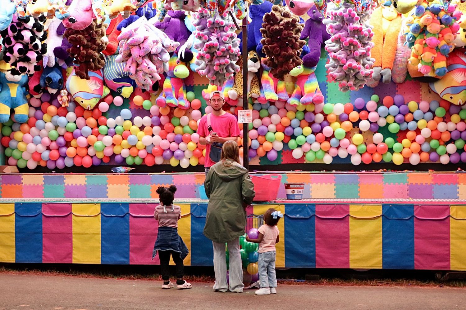 Children and parent at a colorful carnival game booth with balloons and plush prizes.