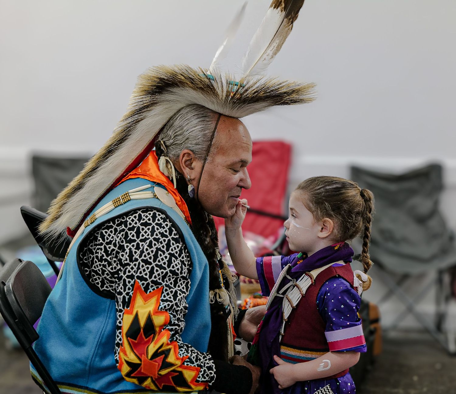 Native American man in traditional regalia with feather headdress helping a young girl with face paint at a cultural event.