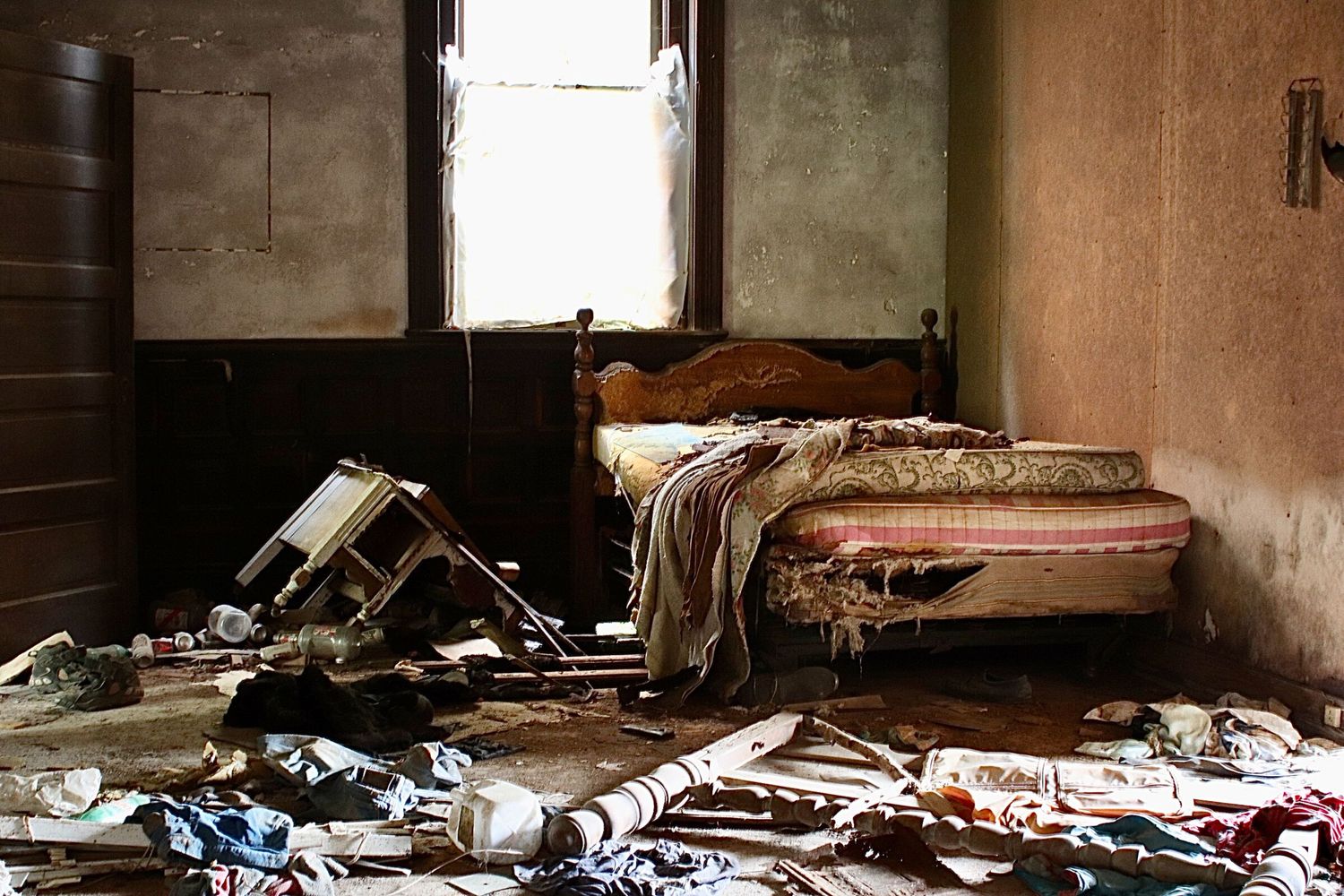 Abandoned bedroom with a torn mattress, scattered belongings, and light coming through a window in a rural home.