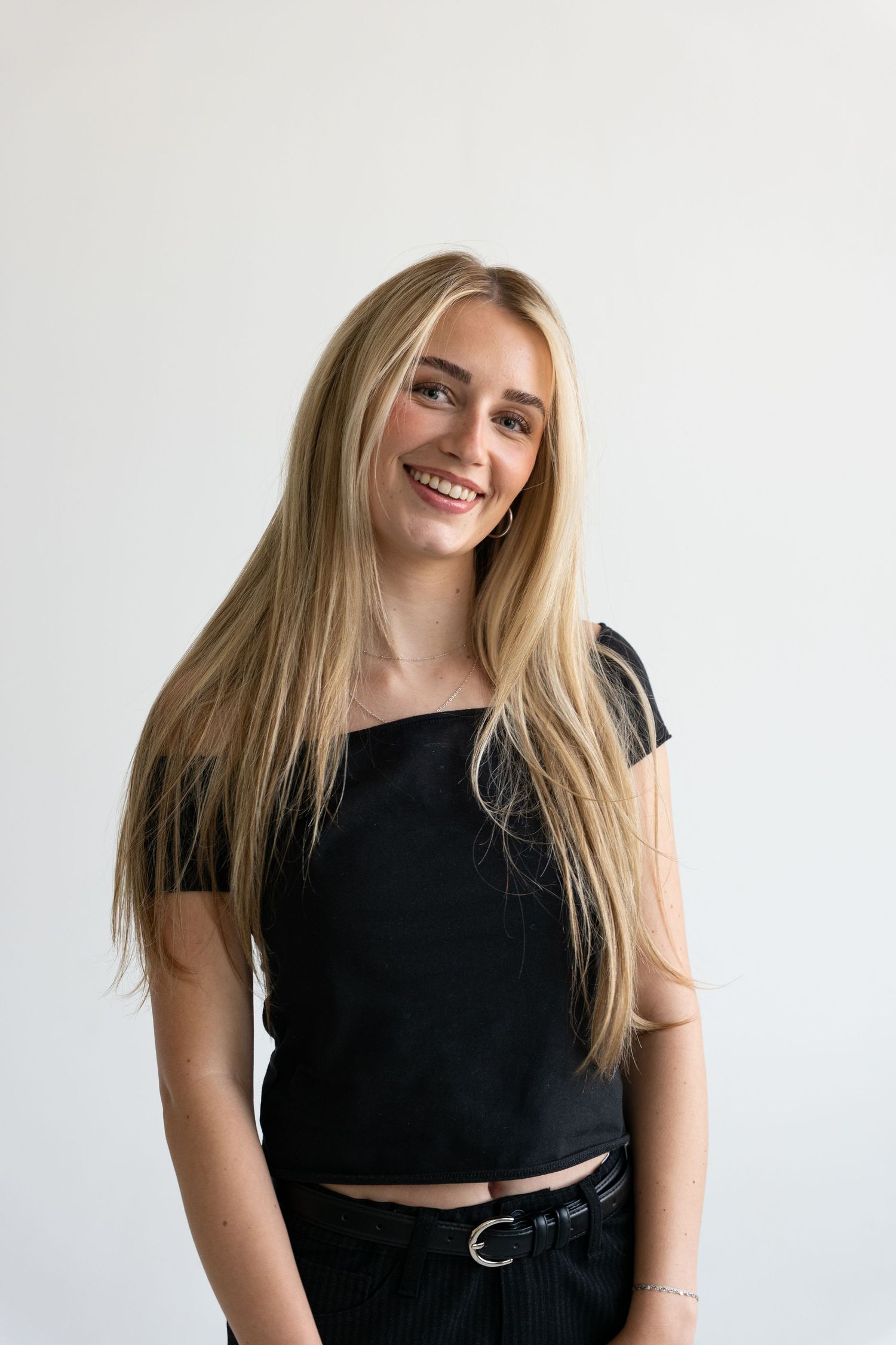 Smiling blonde woman in a black crop top and dark pants posing in a bright white studio.