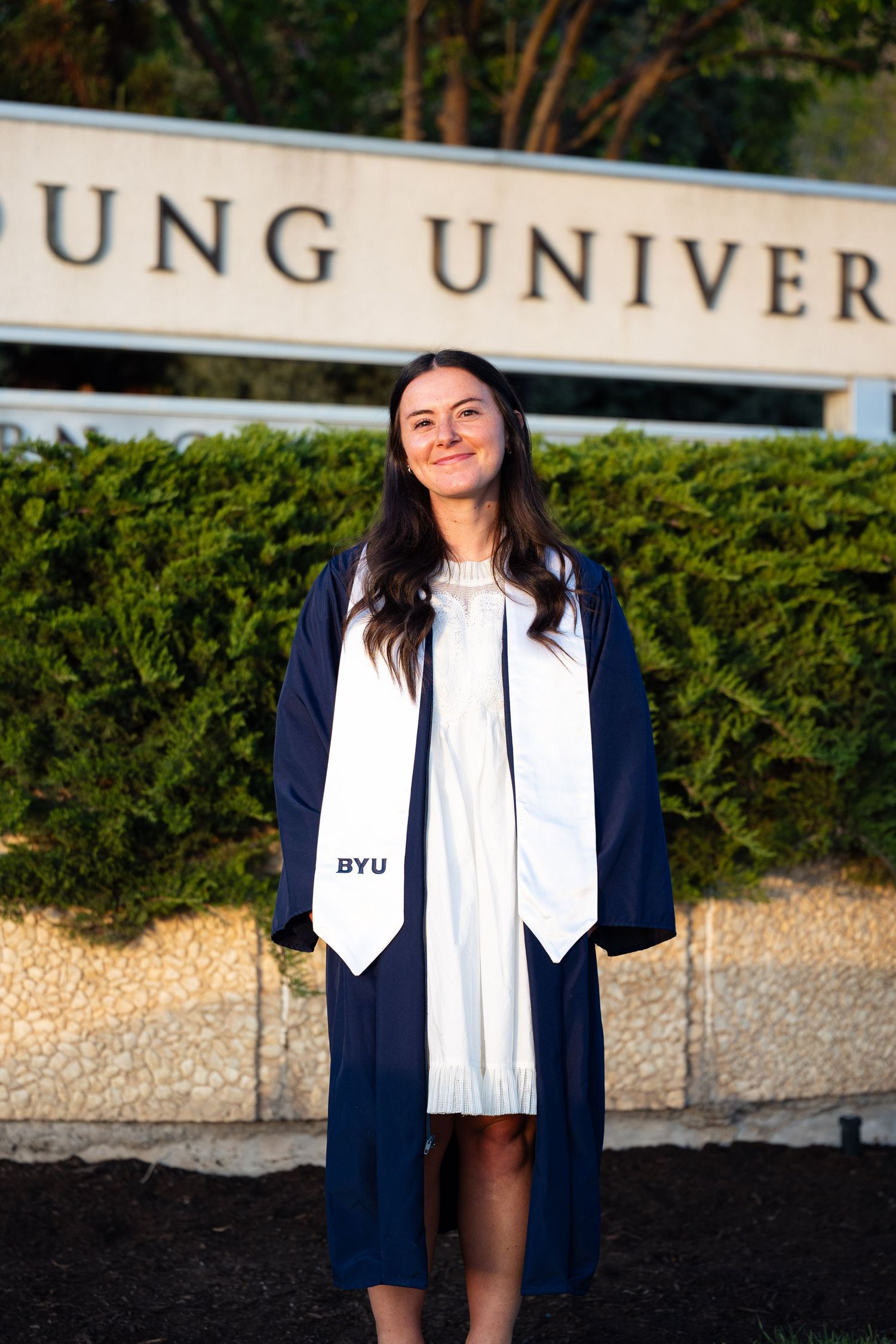 BYU graduate in navy cap and gown with white stole poses in front of Brigham Young University sign.