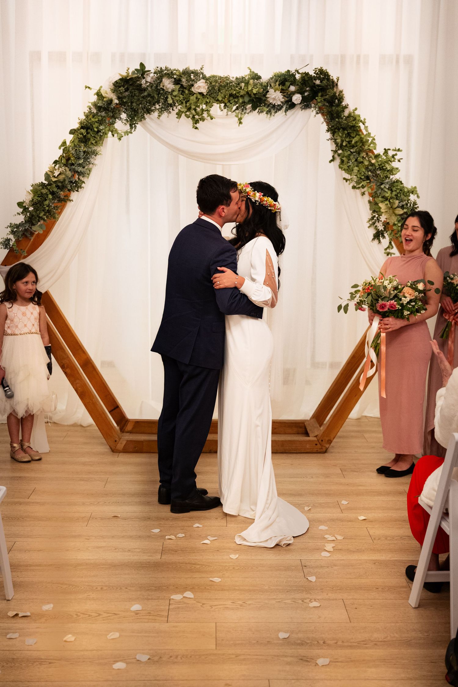 Bride and groom sharing first kiss under hexagonal floral arch at indoor wedding ceremony.