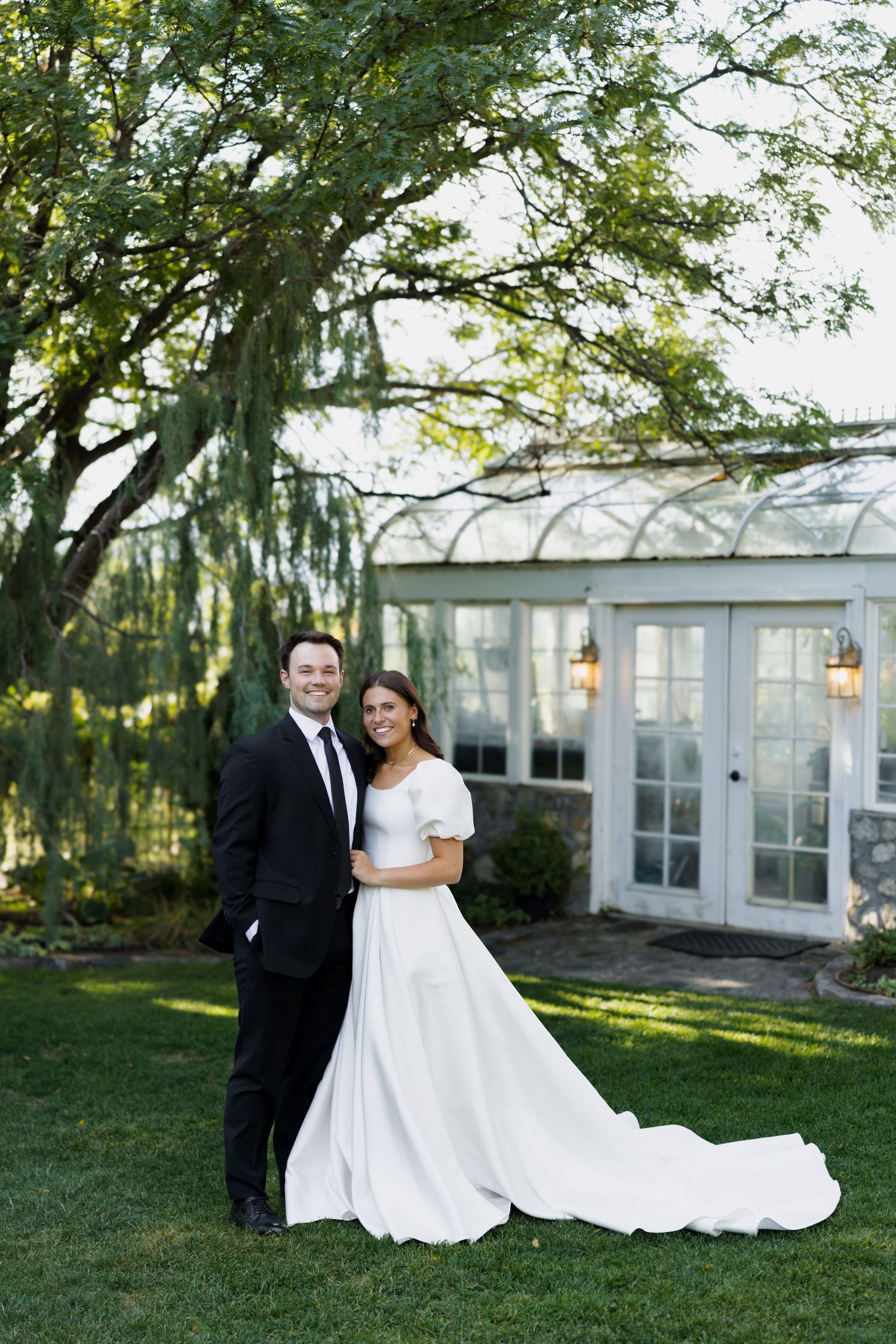 A smiling bride in a white gown and groom in a black suit pose together on a lush green lawn near a greenhouse.