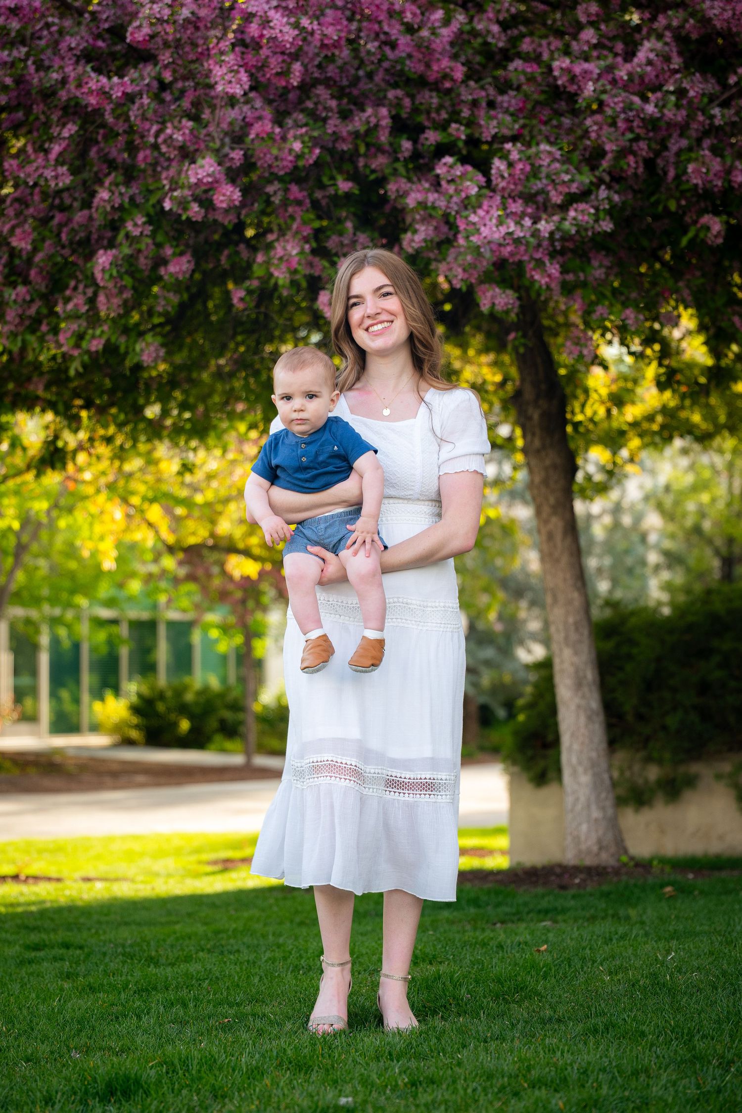A woman in a white dress holds a baby boy in a blue outfit, standing in a garden setting.