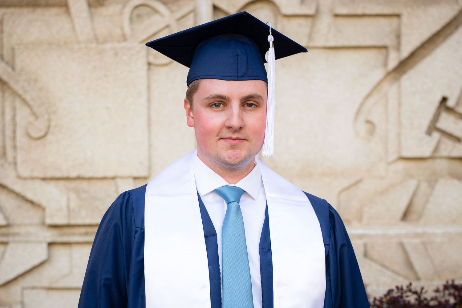 A smiling man in a graduation gown and cap stands in front of a brick building.