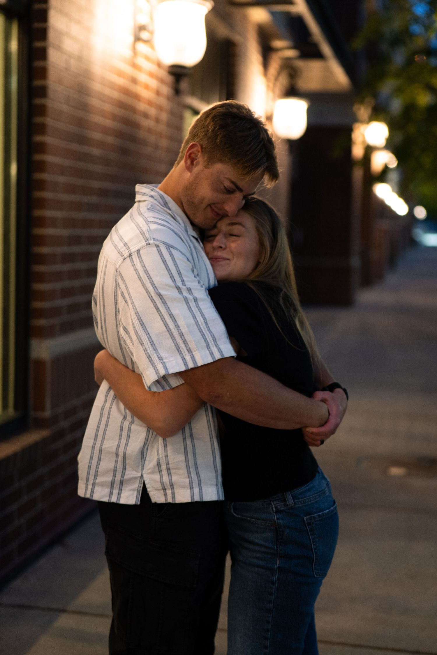 Couple embracing tenderly on a city sidewalk at dusk, warm lights glowing behind them near a brick building.