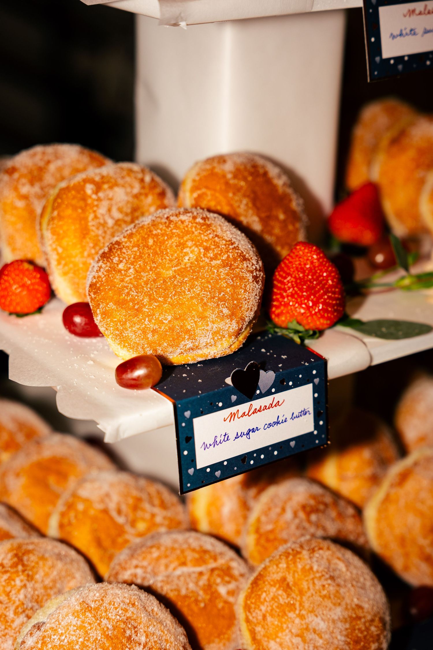 Sugar-dusted round pastries displayed on tiered trays with fresh strawberries and a decorative label at a wedding.
