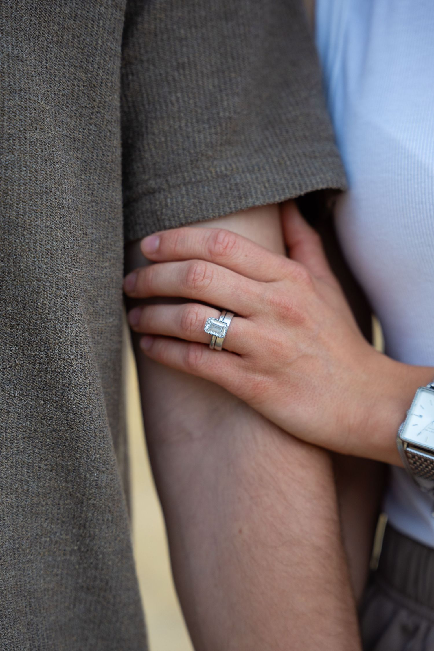 Close-up of a woman's hand wearing a diamond engagement ring and silver watch, gently holding a man's arm.