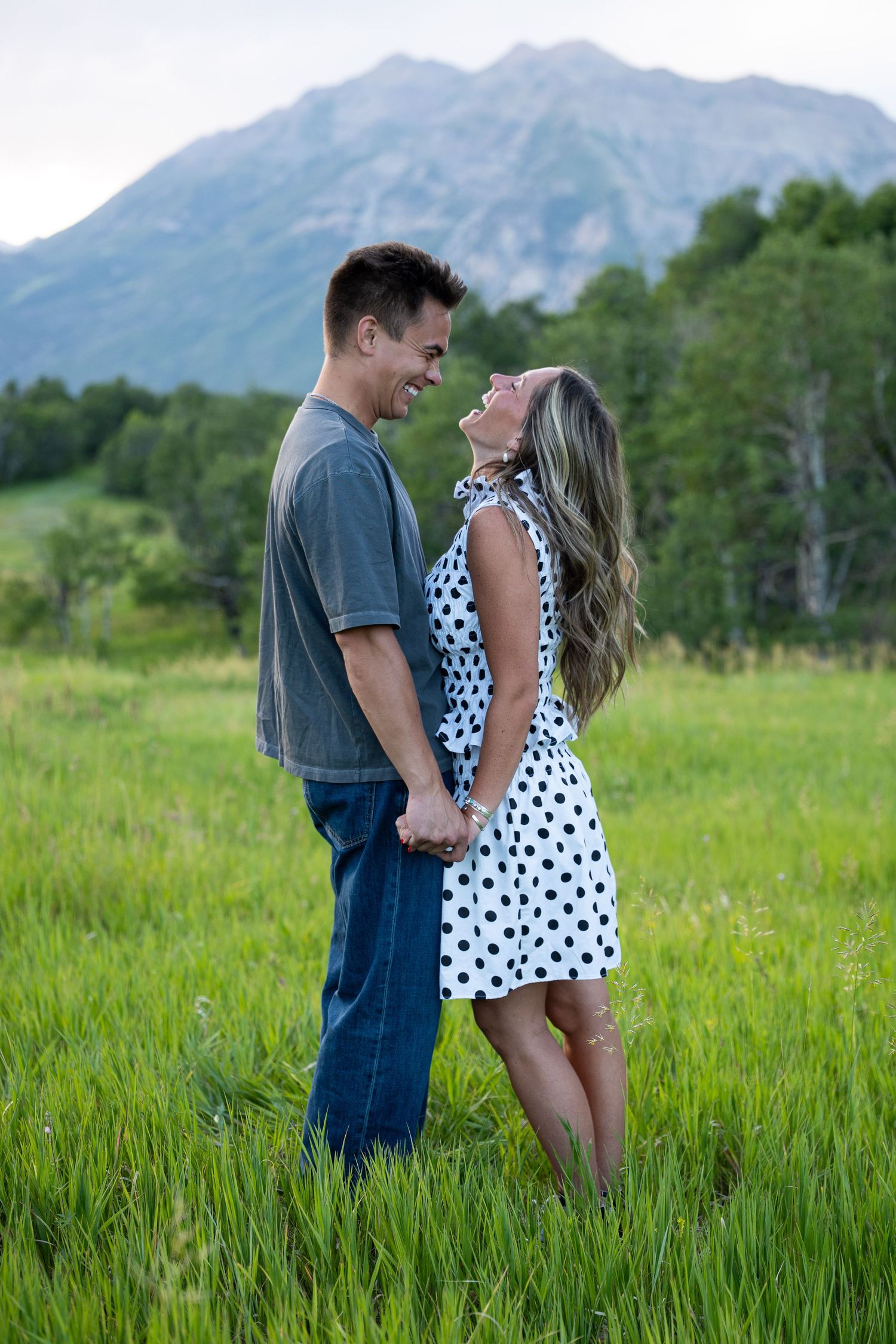 Happy couple holding hands in a green meadow with mountain backdrop, woman in polka dot dress laughing.