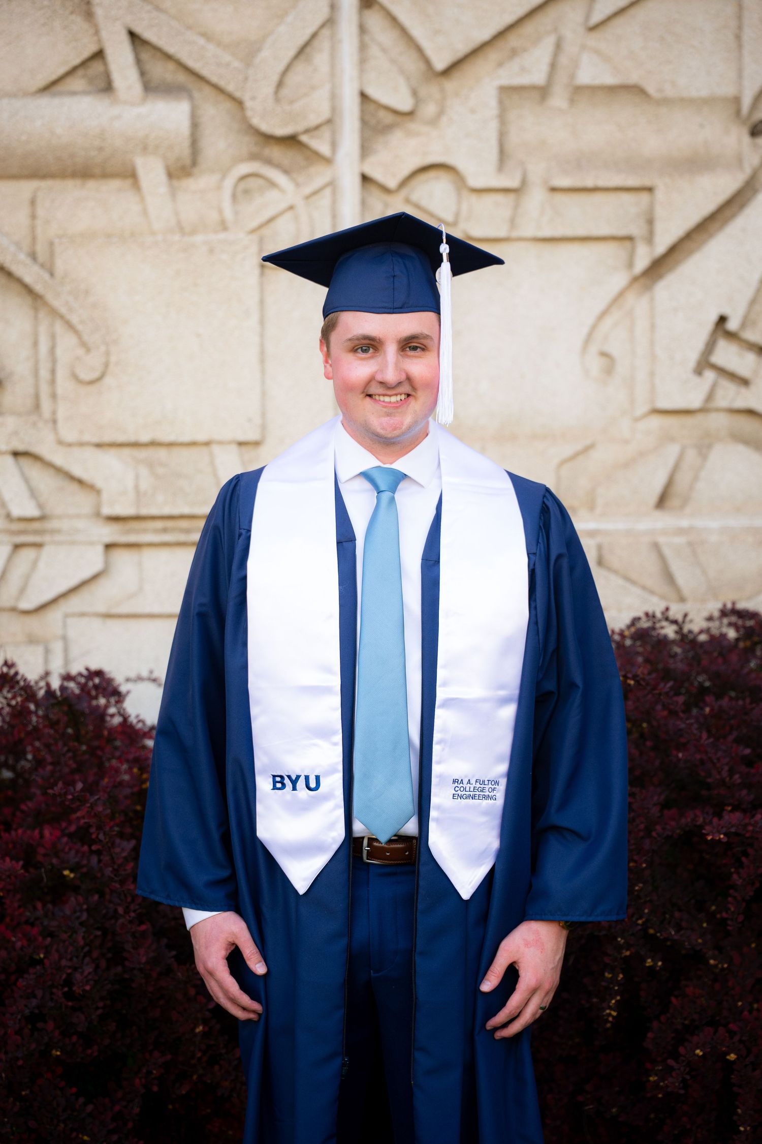 A man in a graduation gown and cap stands in front of a university building.