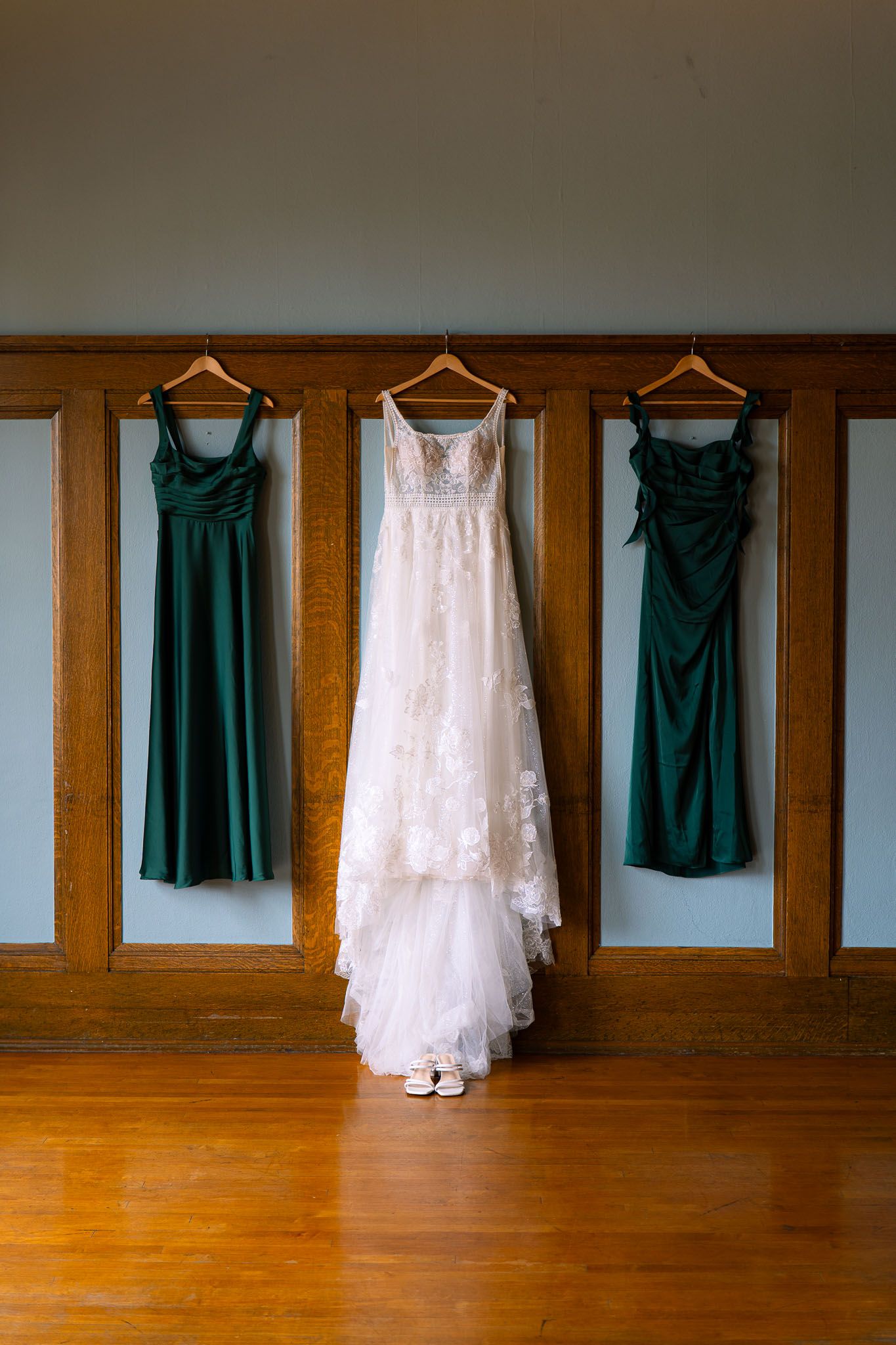 White lace wedding gown flanked by two emerald green bridesmaid dresses hanging on wooden wall panels with bridal shoes below.