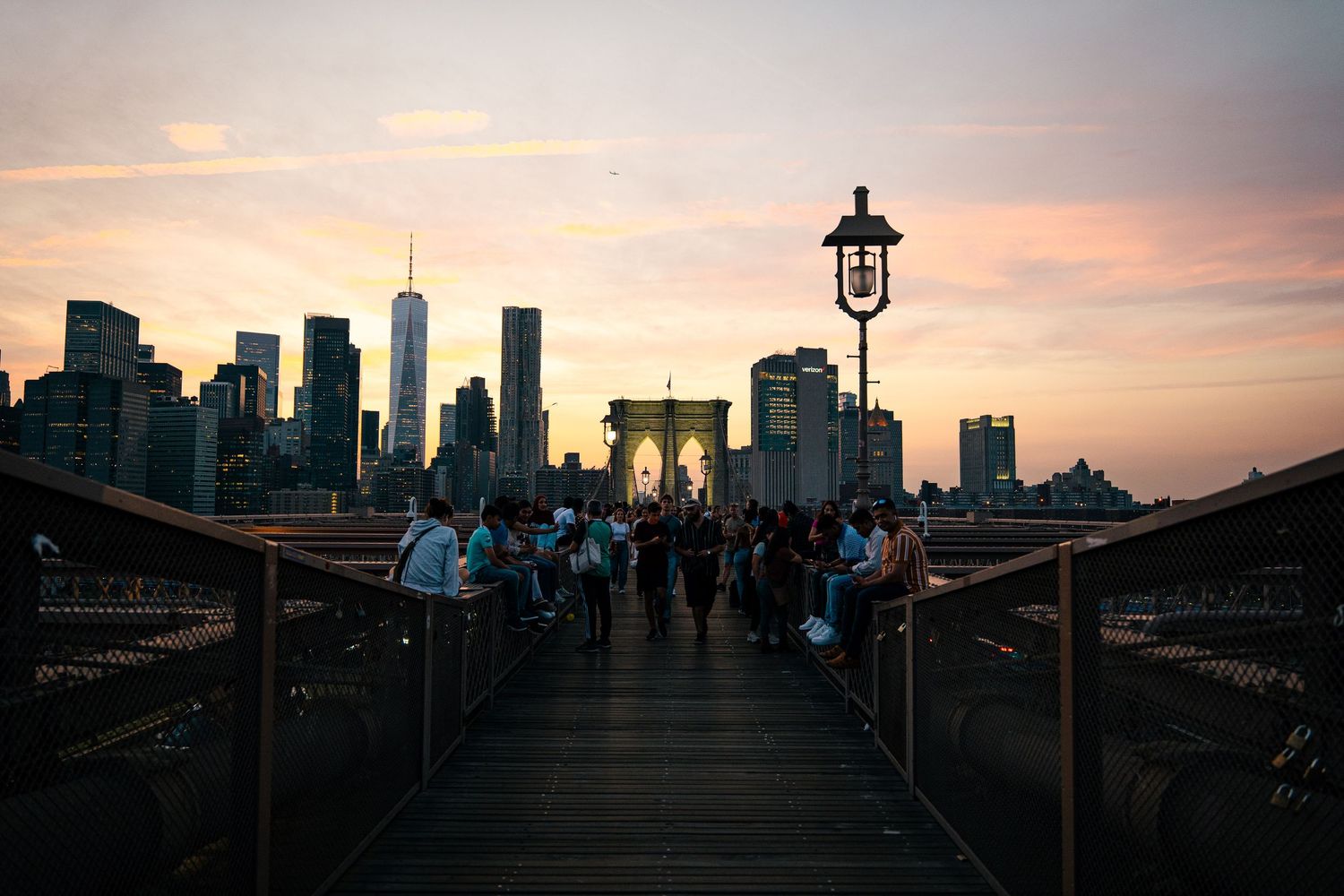 Pedestrians walking on the Brooklyn Bridge at sunset, with the Manhattan skyline in the distance.