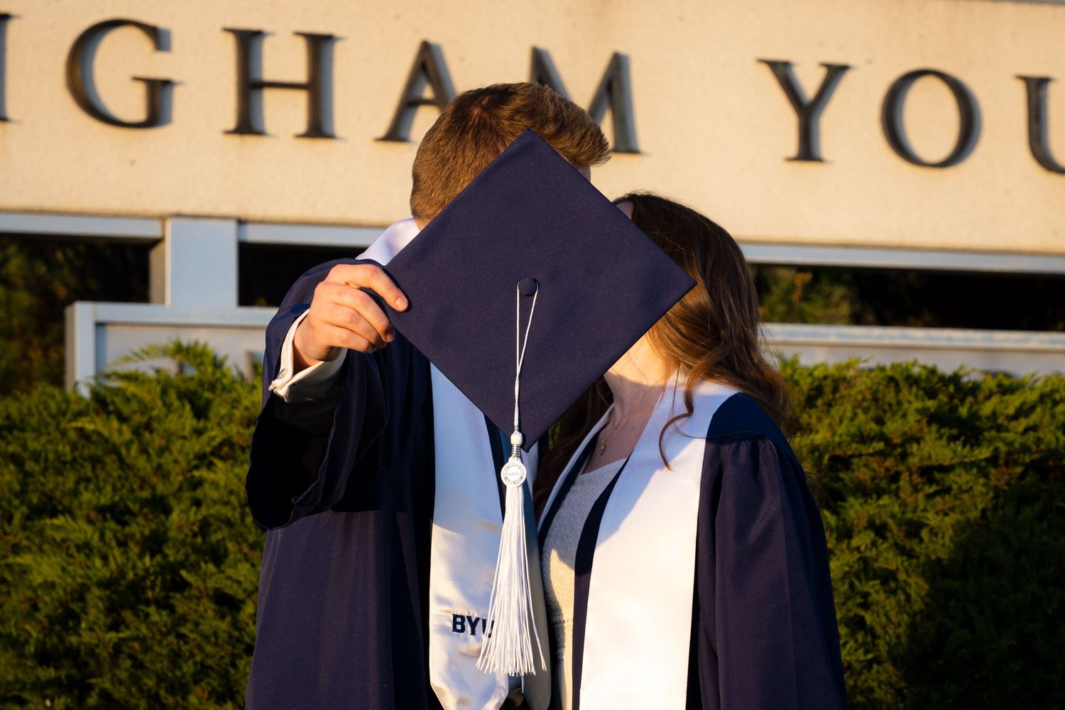 A young woman with long curly brown hair smiles while wearing a white dress and blue graduation gown in front of a building.