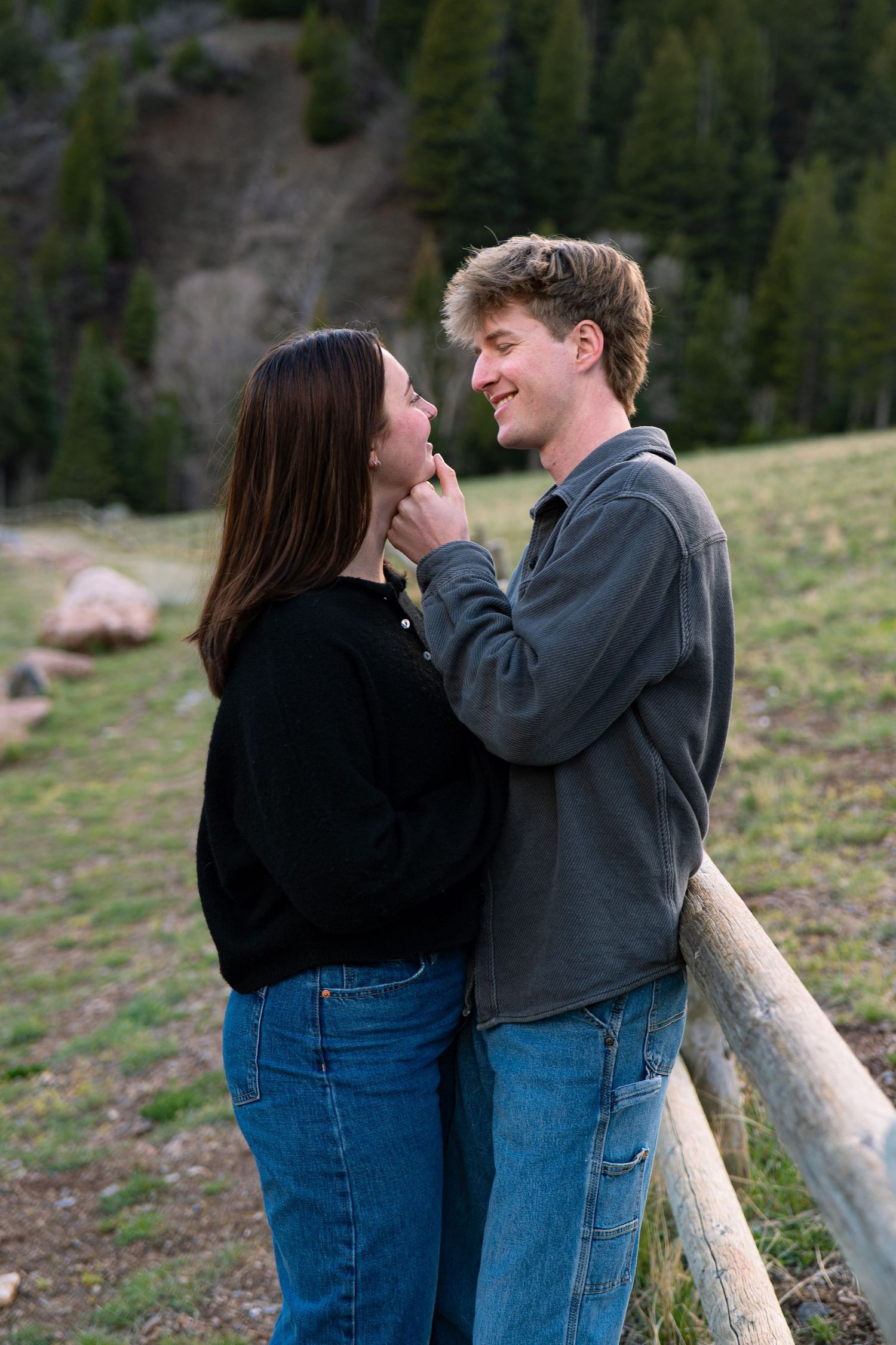 Young couple sharing a romantic moment by a wooden fence with mountain forest backdrop.