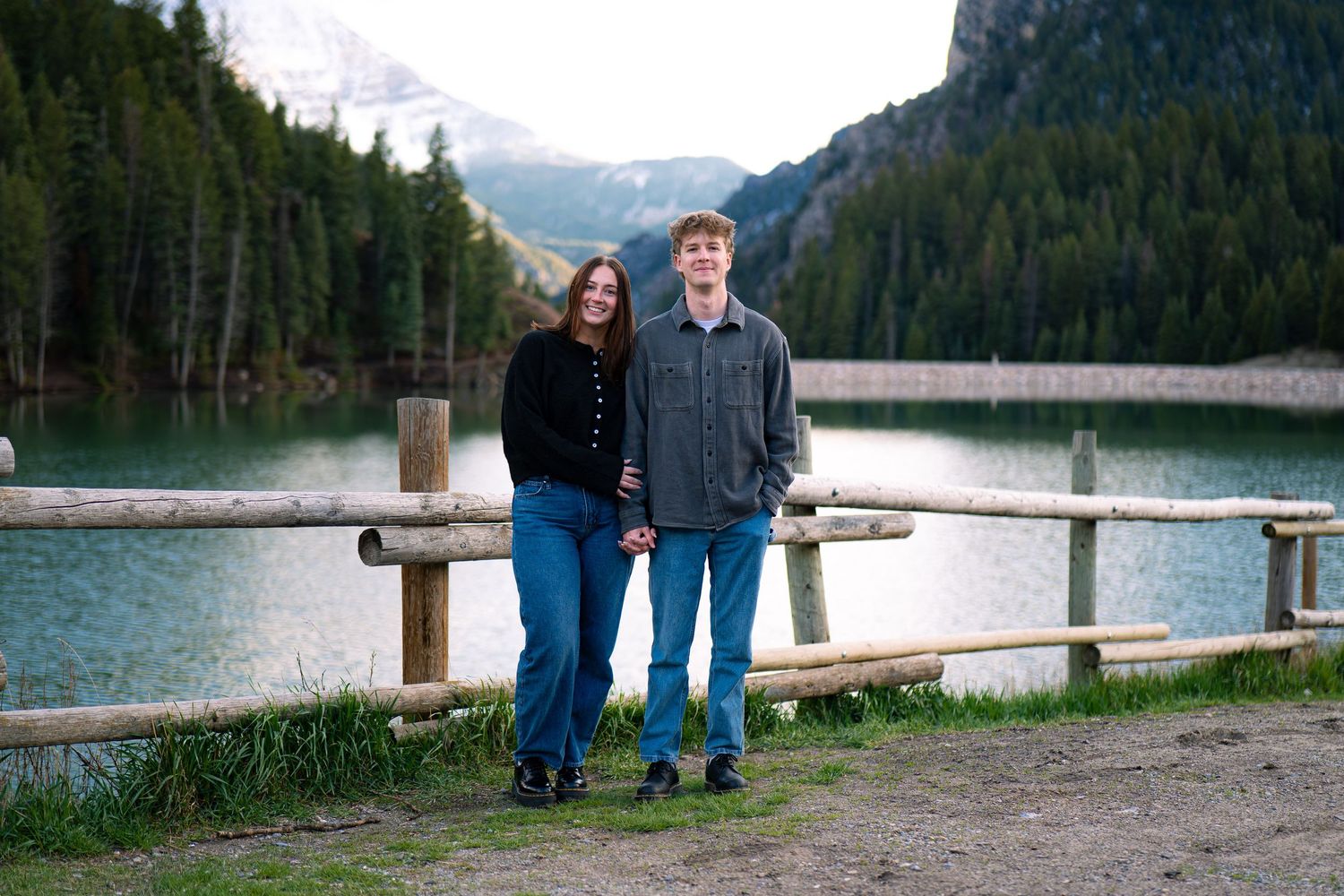 Couple posing together by a wooden fence at a mountain lake surrounded by evergreen forest.