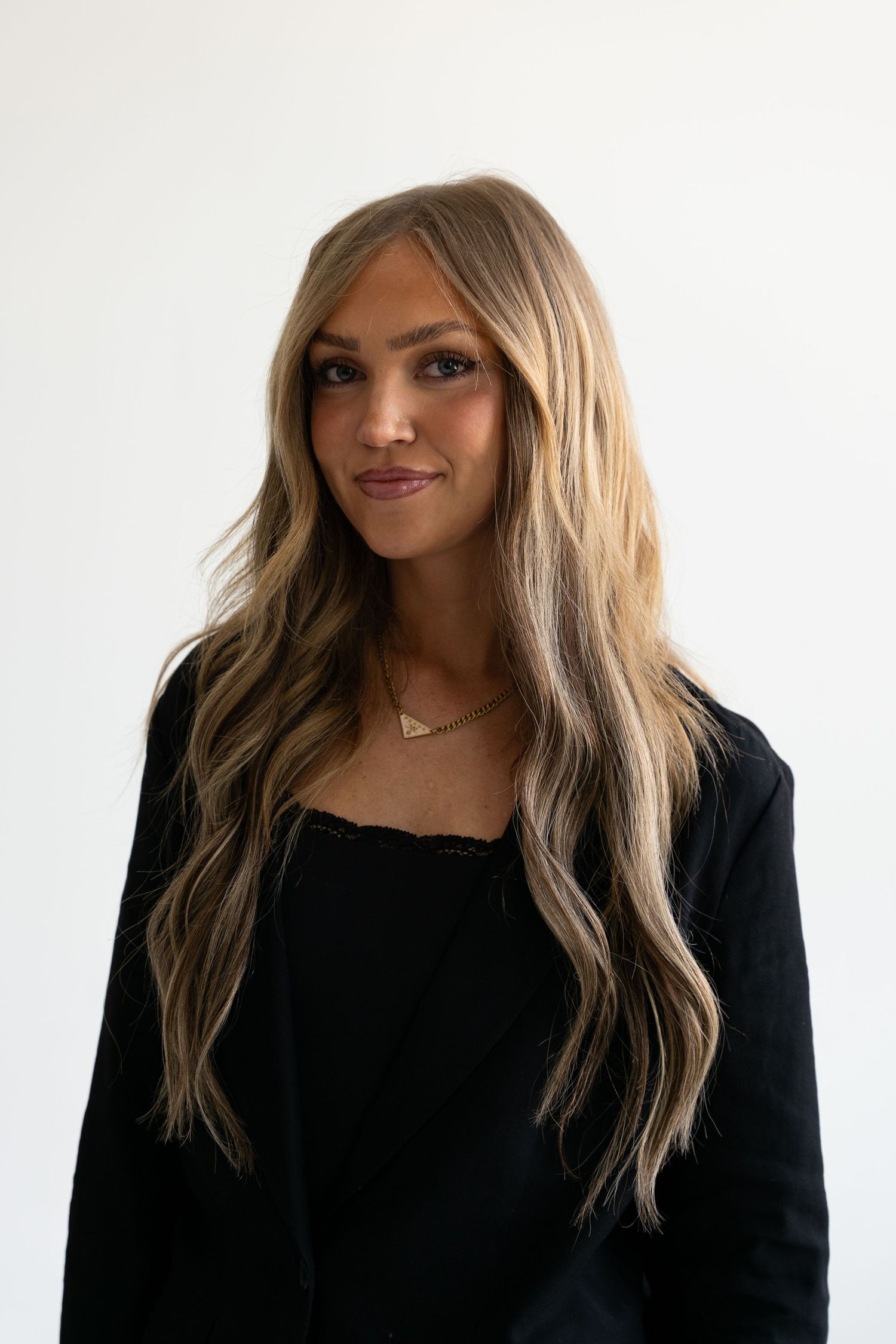 Blonde woman with long wavy hair wearing a black top and gold necklace posing in a white studio.