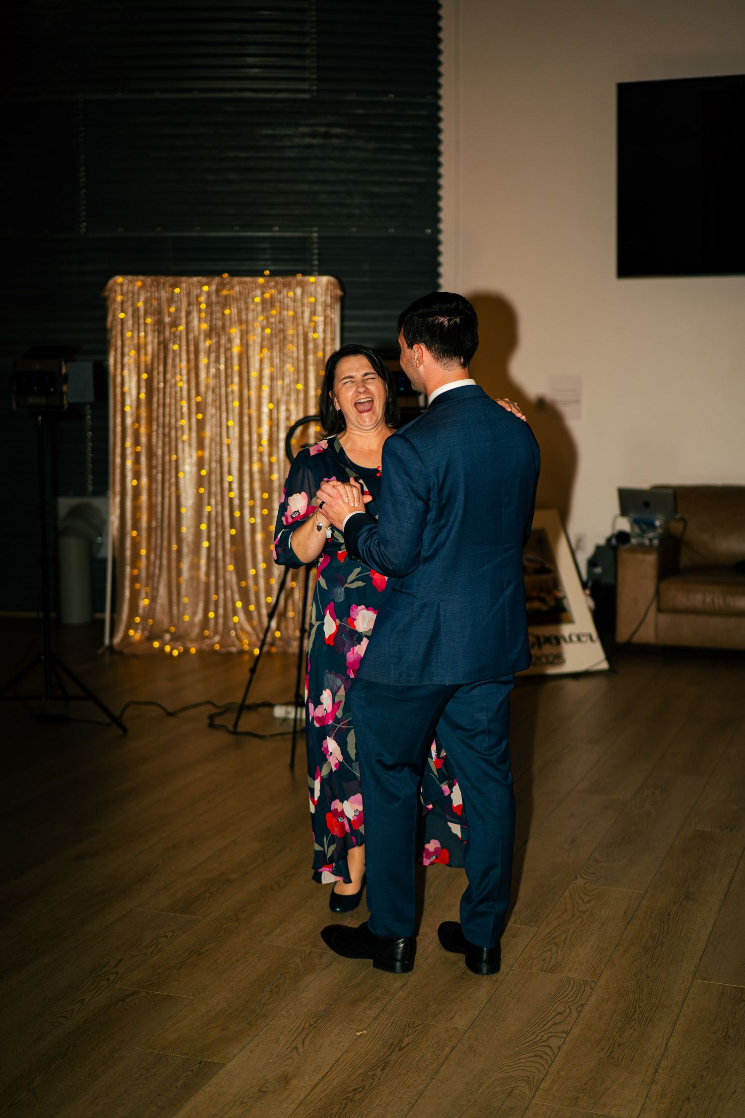 Man in navy suit dancing with woman in floral dress at wedding reception with gold sequin backdrop.
