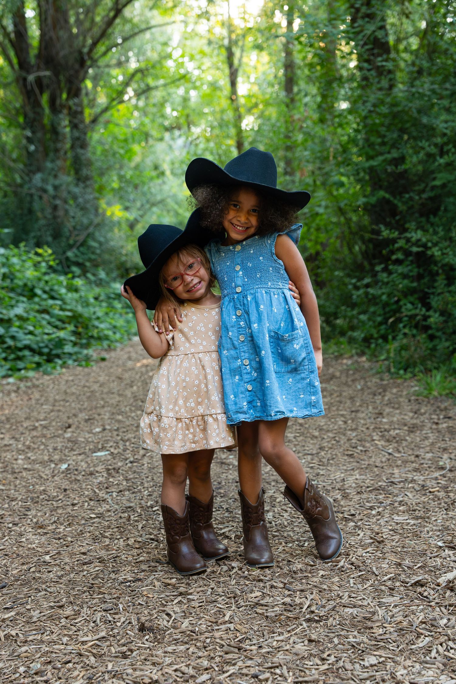 Two young girls in cowboy hats, dresses, and boots smile and hug on a woodland path.