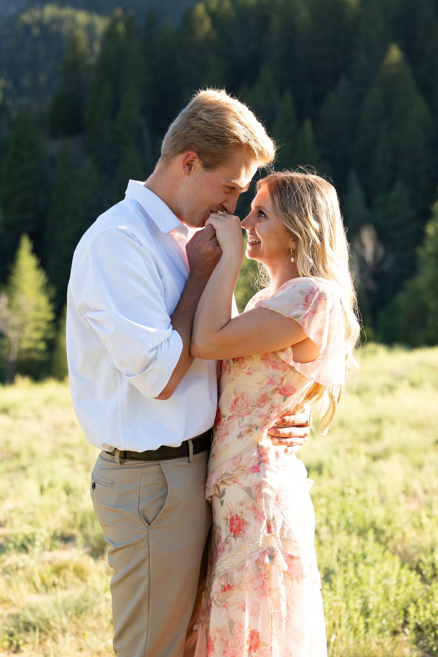 Couple embracing in a sunlit meadow, man in white shirt holding woman in floral dress with pine trees behind.