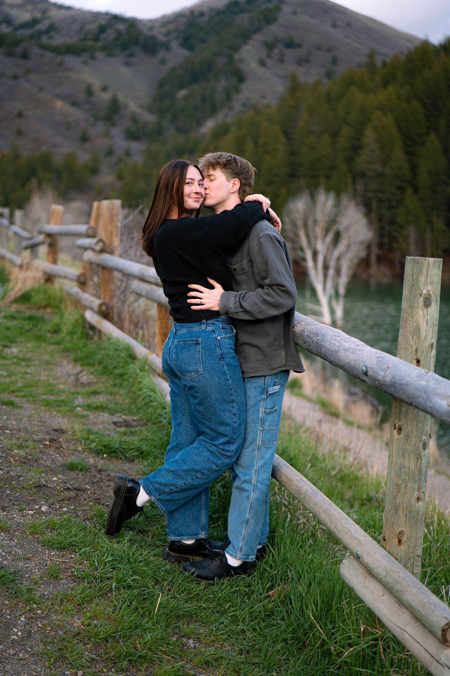 A couple embraces and kisses by a rustic wooden fence with green hills and mountains in the background.
