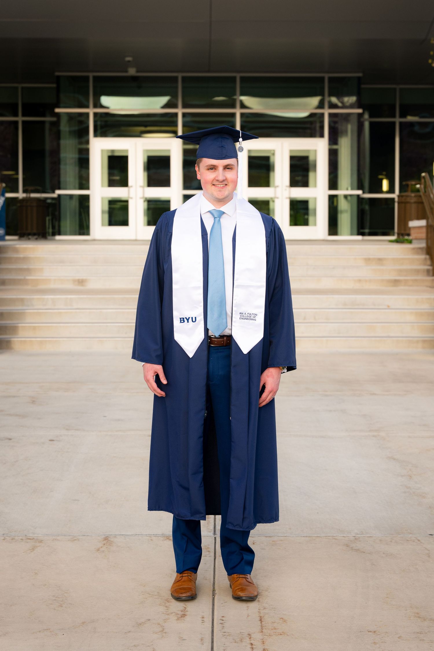 A young man wearing a blue graduation gown and cap stands in front of a building.
