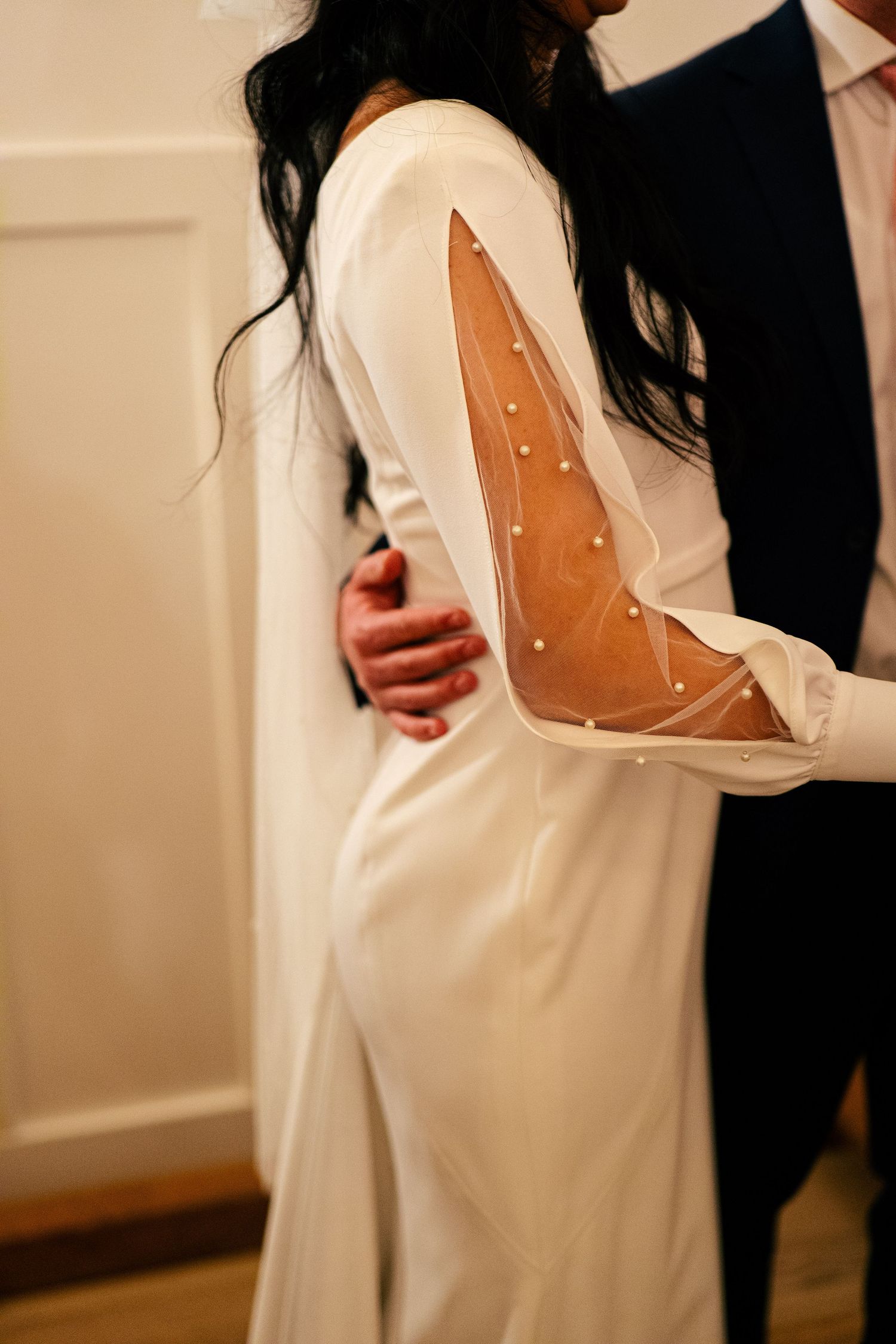 Bride in elegant white long-sleeve wedding gown with sheer pearl-embellished back detail embraced by groom.