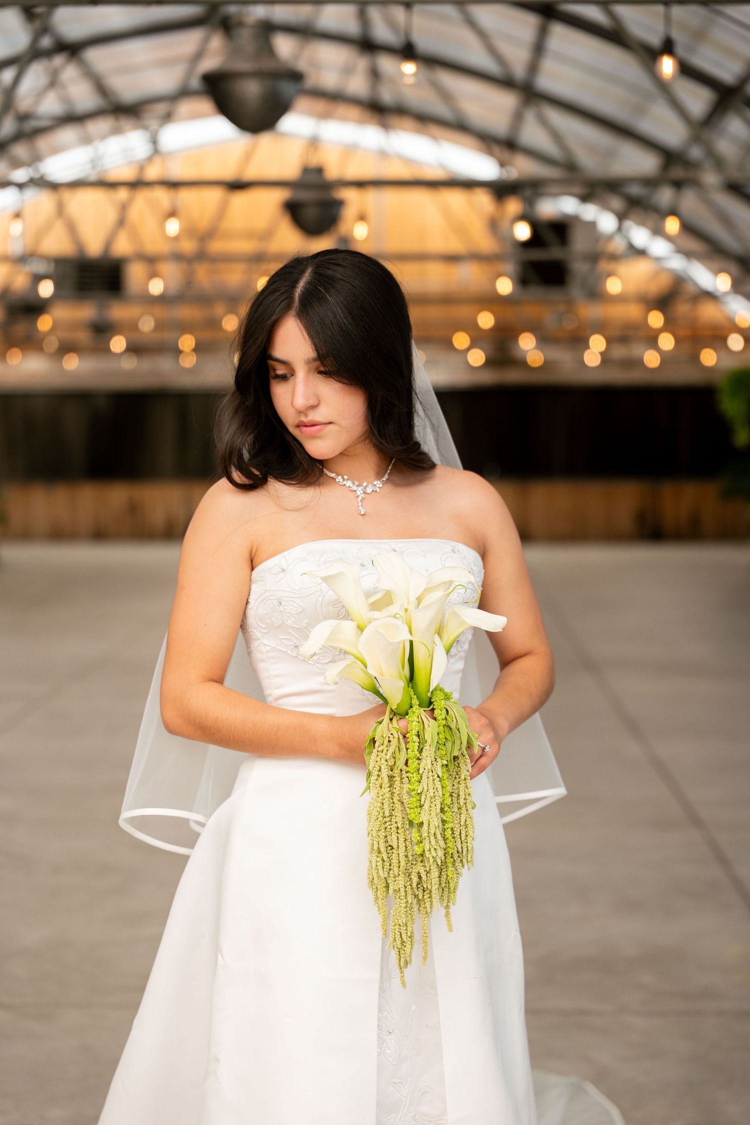 Bride in strapless white gown holding calla lily bouquet with green accents, standing indoors with warm bokeh lights behind her.