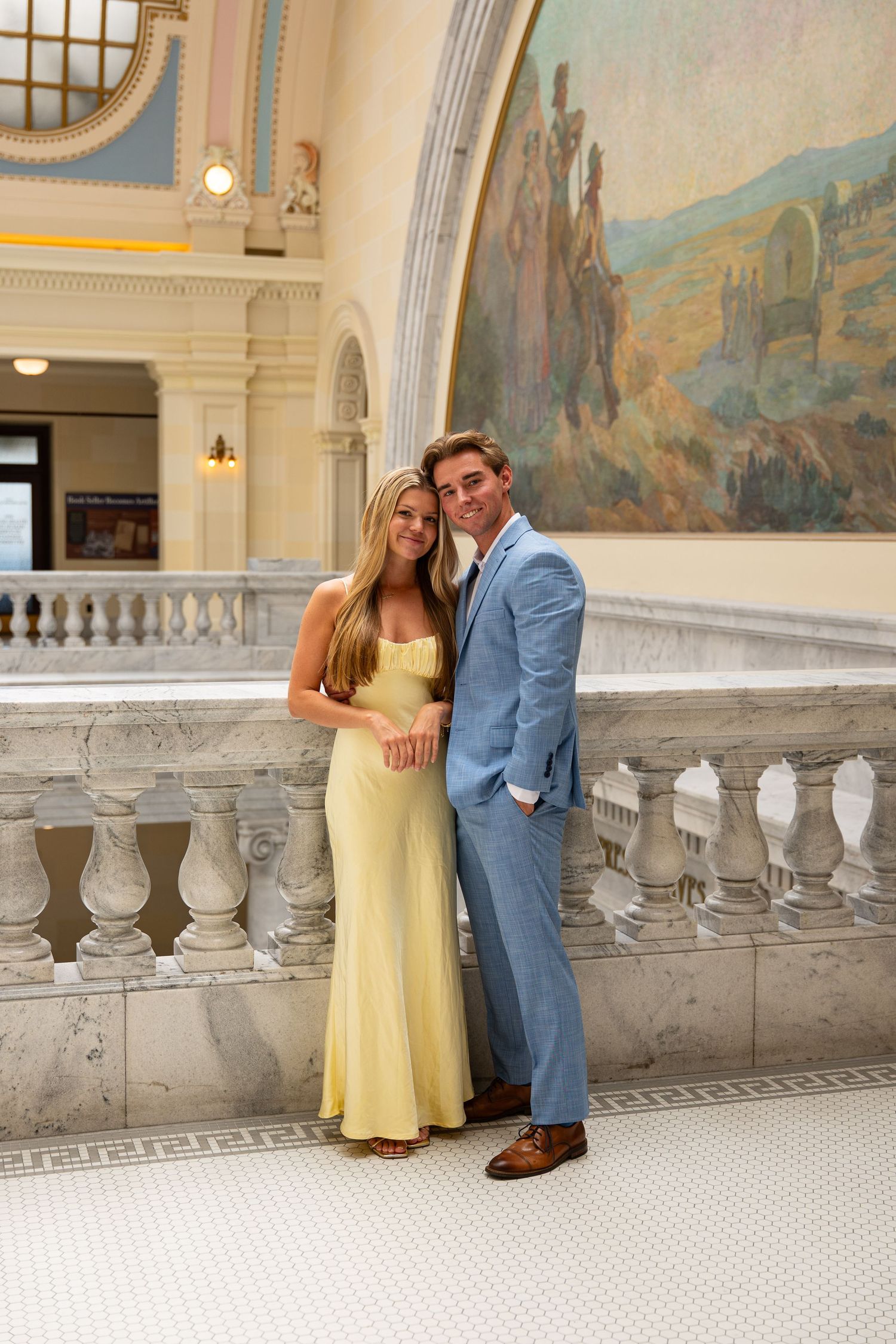Couple in formal attire posing by marble balustrade in elegant building with classical mural backdrop.