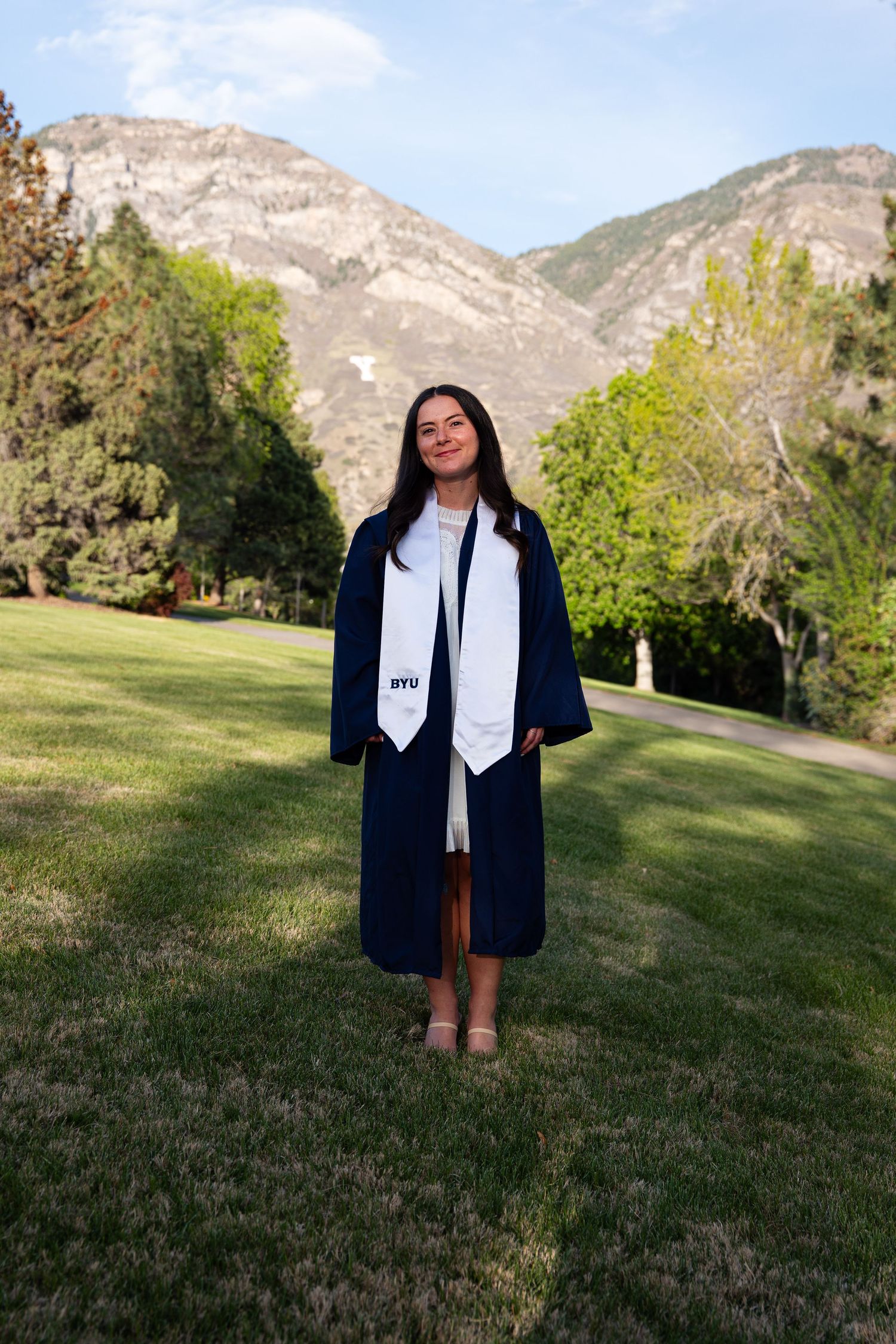 BYU graduate in navy cap and gown with white stole stands on green lawn with mountain backdrop.