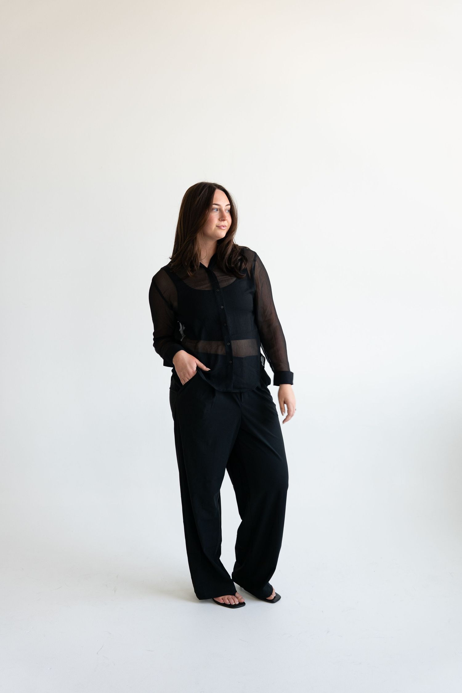 Brunette woman in black sheer button-up top and wide-leg trousers posing in a minimalist white studio.
