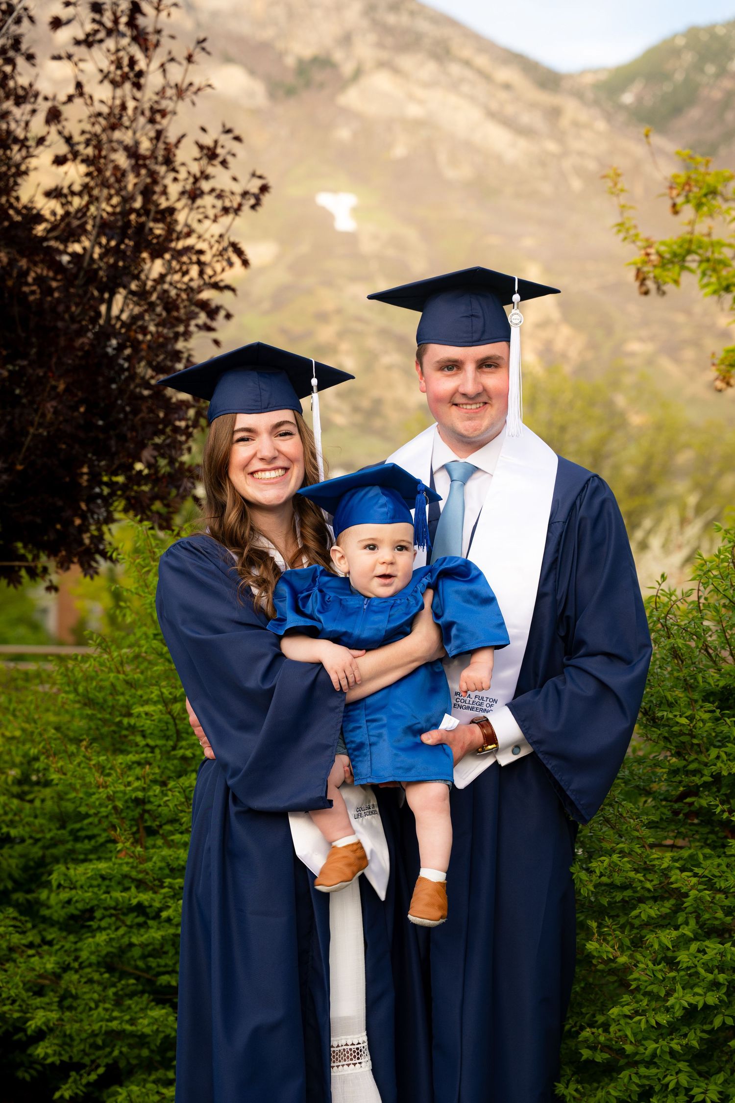 A family of three, including a baby, pose for a portrait outdoors.