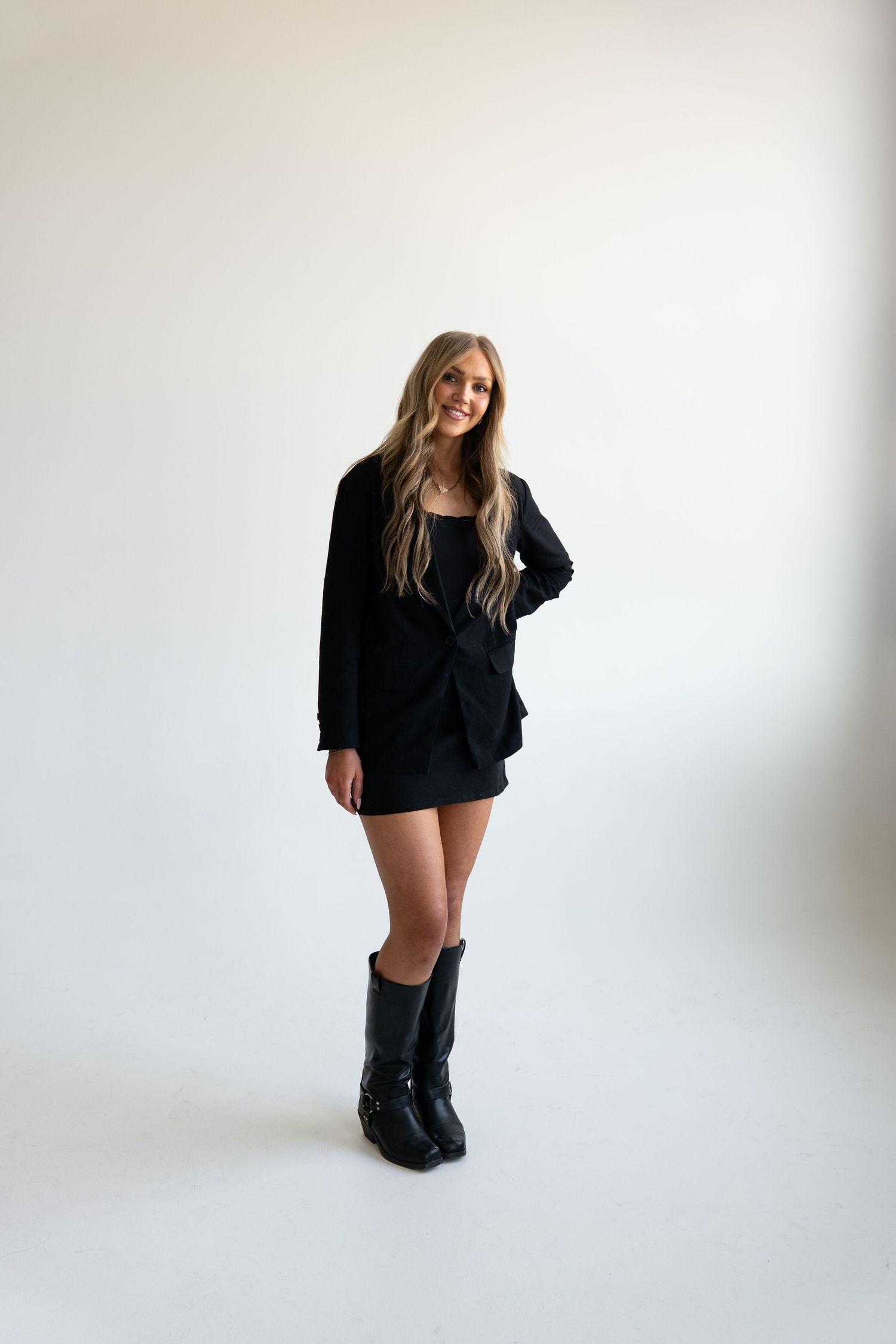 Blonde woman in black mini dress and knee-high boots on white background.